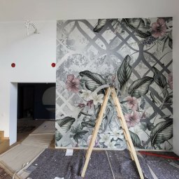 Adrian Zaliwski - Wnętrze w trakcie dekoracji: ściana z motywem roślinnym, drabina oparta o ścianę, zabezpieczona podłoga. Widoczny fragment schodów i przejście do innego pomieszczenia.