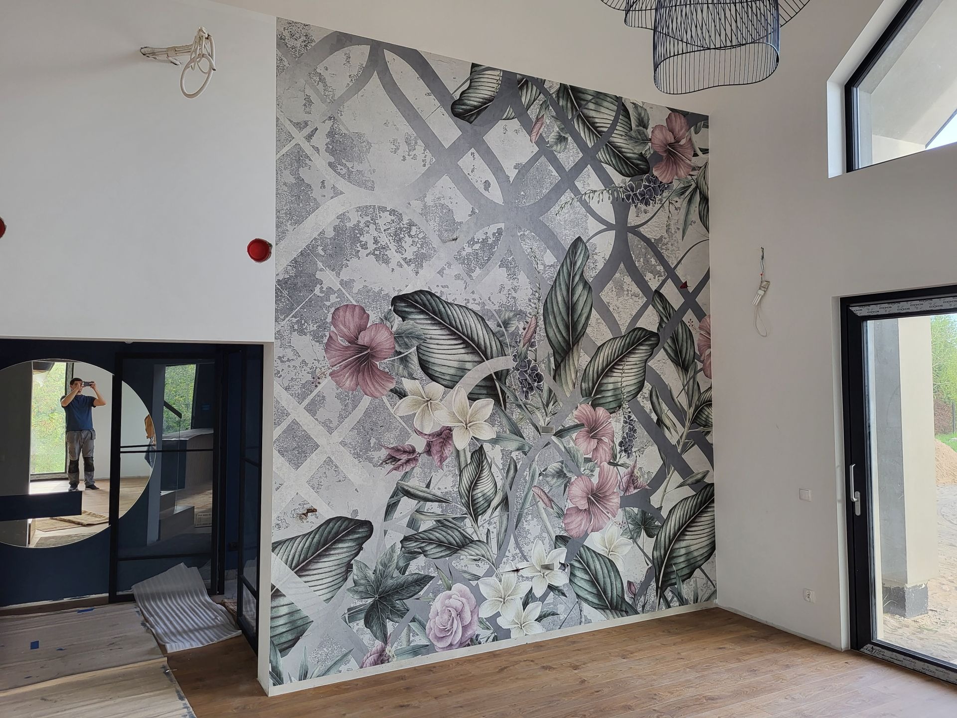 Wnętrze z dekoracyjną tapetą w kwiaty i liście na ścianie, drewniana podłoga. Osoba robiąca zdjęcie w lustrze. Nowoczesna lampa wisząca.