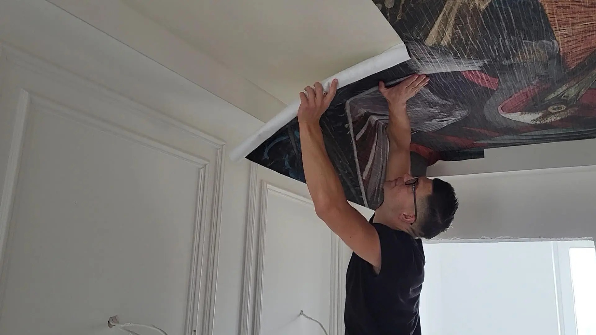 Mężczyzna w okularach nakleja fototapetę na sufit w pomieszczeniu z białymi ścianami i ozdobnymi listwami. Widoczne ręce i fragment wzoru fototapety.