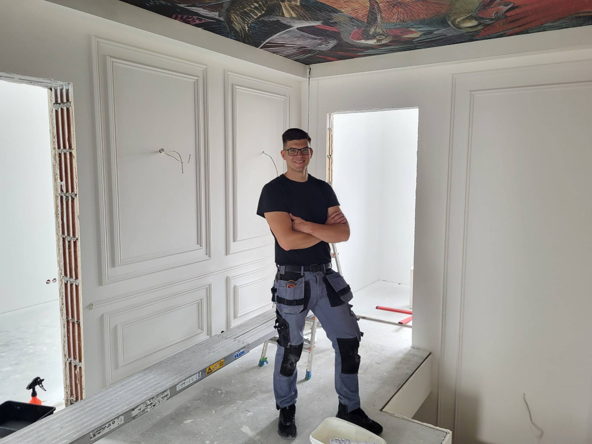 Malarz w roboczym stroju na tle białych ścian z dekoracyjnymi panelami i malowidłem na suficie. Widoczny drabina i narzędzia malarskie.