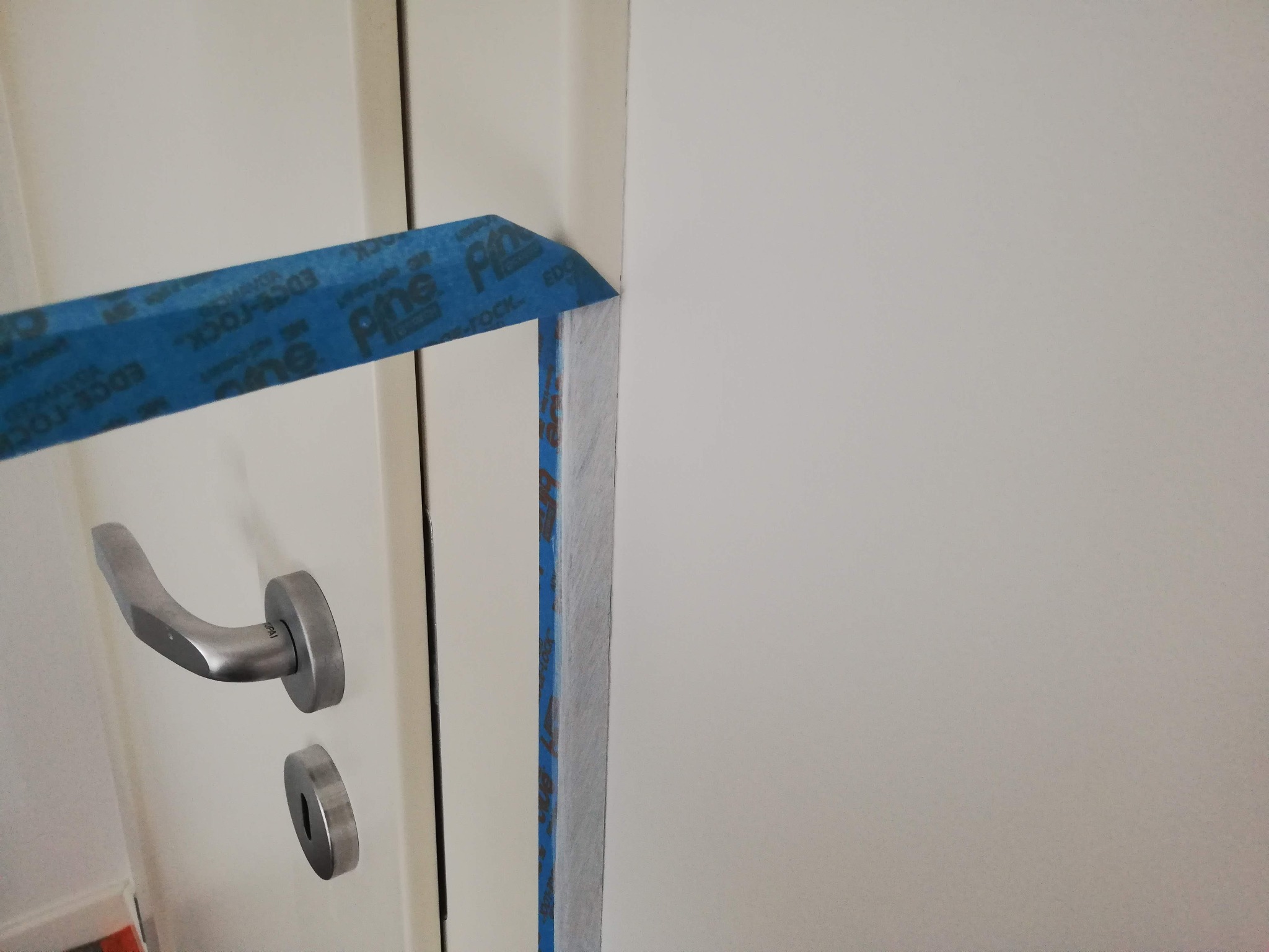 Przygotowanie do odświeżania framugi drzwi: niebieska taśma malarska zabezpiecza krawędzie przed aplikacją farby na białe drzwi z metalową klamką.