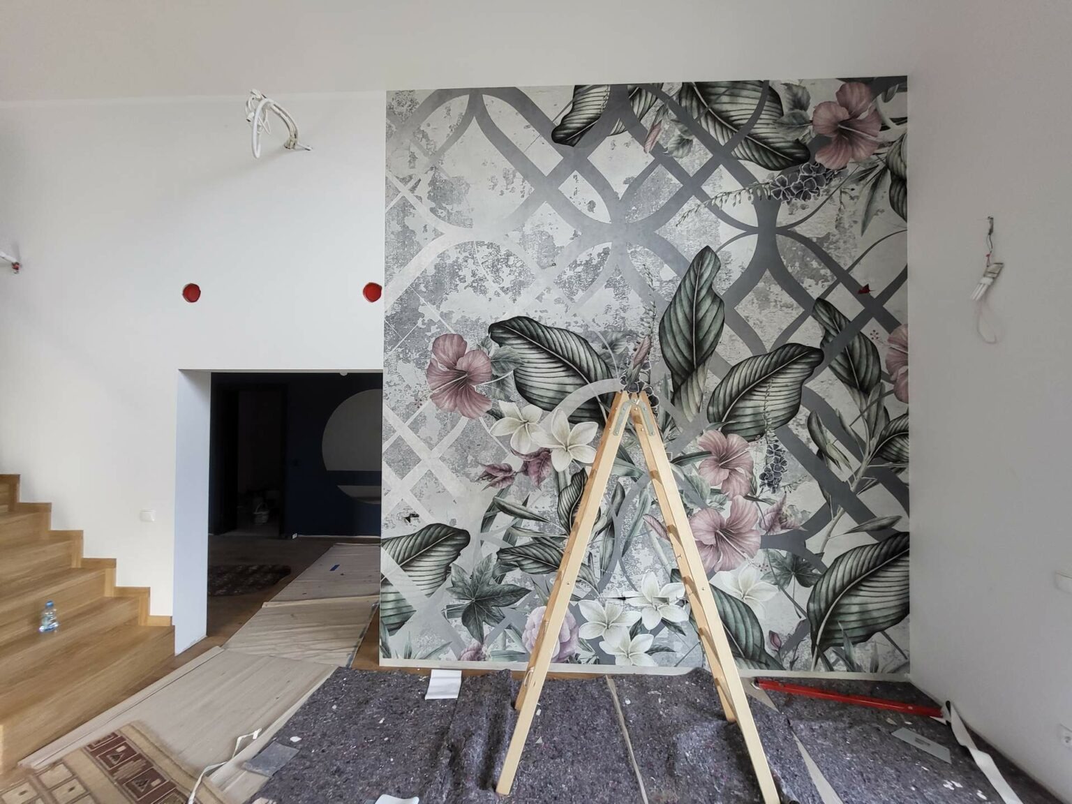 Wnętrze w trakcie dekoracji: ściana z motywem roślinnym, drabina oparta o ścianę, zabezpieczona podłoga. Widoczny fragment schodów i przejście do innego pomieszczenia.