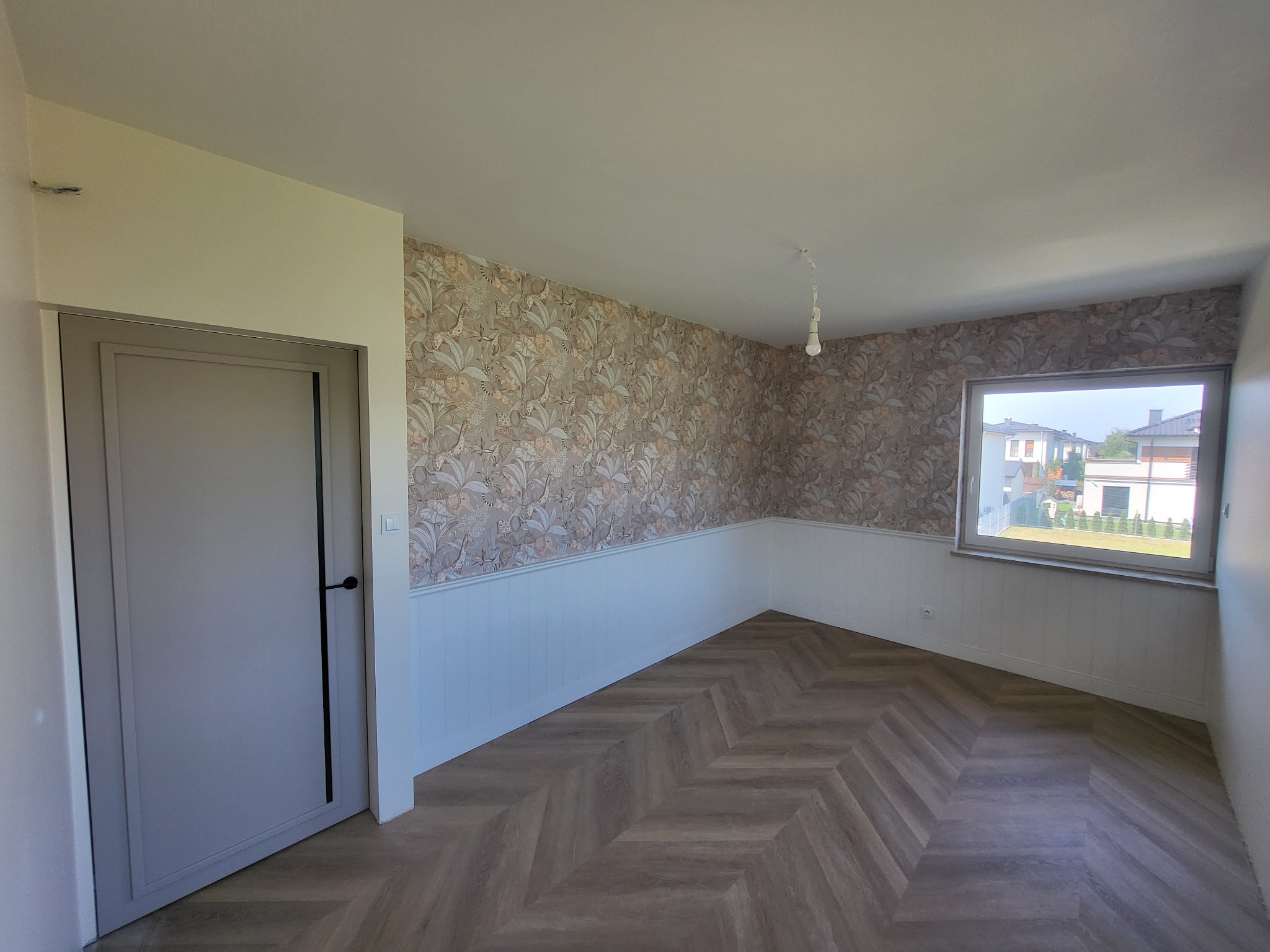 Pokój po remoncie: wzorzysta tapeta w liście, biała lamperia, parkiet w jodełkę, szare drzwi. Widok na dom z ogrodem przez okno.