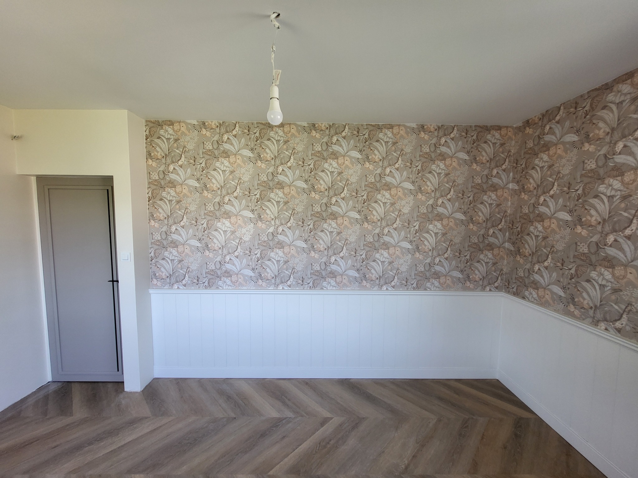 Wnętrze po remoncie: wzorzysta tapeta z motywem roślinnym i zwierzęcym, biała boazeria, podłoga w jodełkę, szara futryna drzwi. Żarówka zwisa z sufitu.