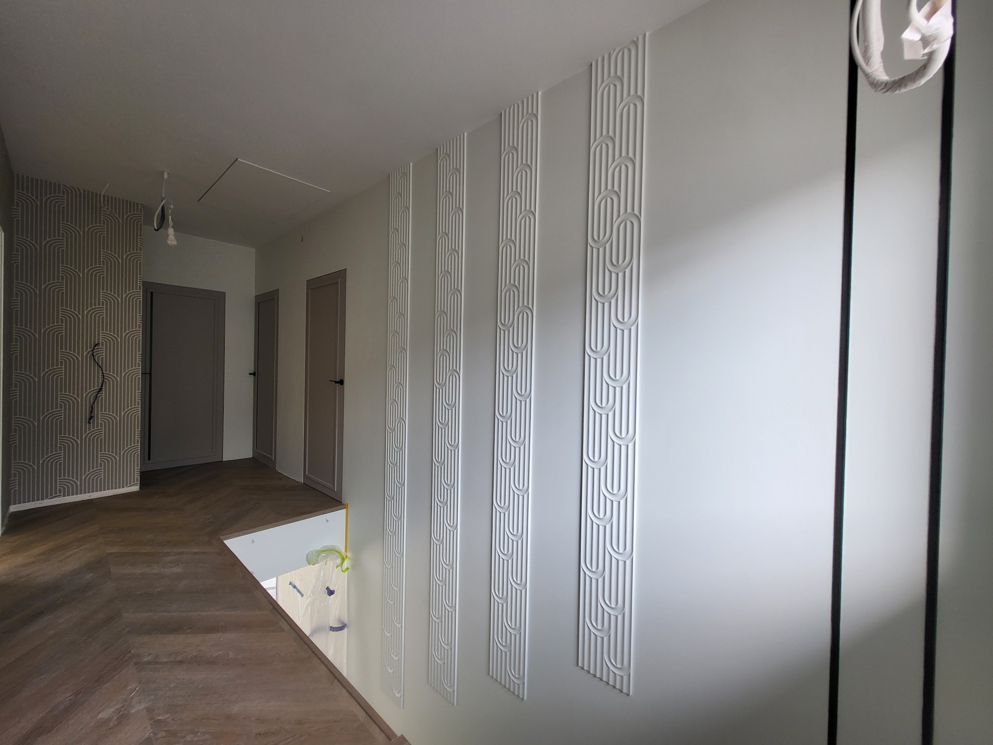 Białe, dekoracyjne panele ścienne z motywem owalnym na jasnej ścianie w korytarzu. Podłoga z parkietu w jodełkę. Widoczne drzwi i tapeta w tle.