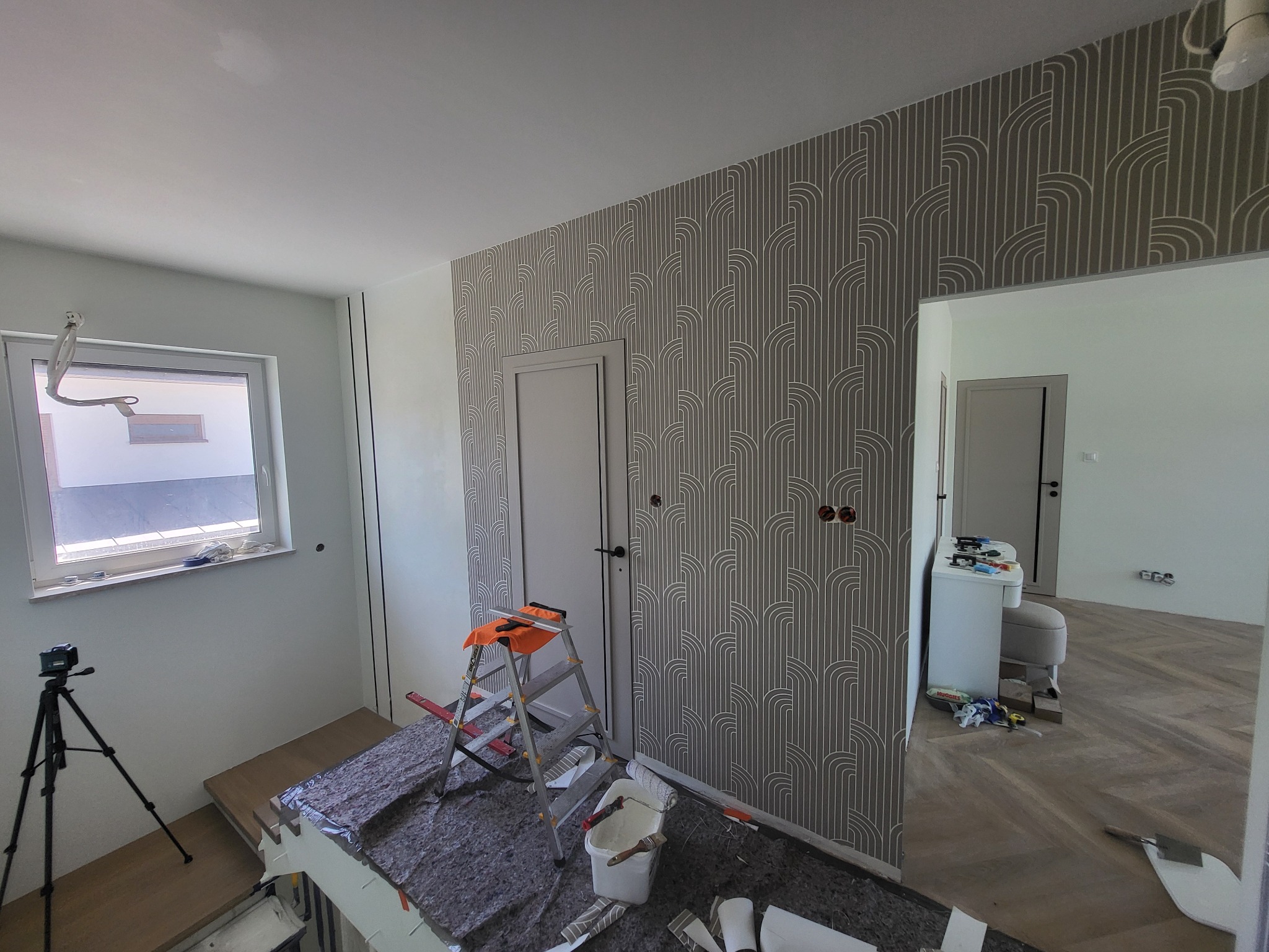 Wnętrze w trakcie prac: ściana z wzorzystą tapetą, drabina malarska z pomarańczową szmatką, wiadro z farbą i wałkiem. Widoczny fragment okna i pomieszczenia obok.