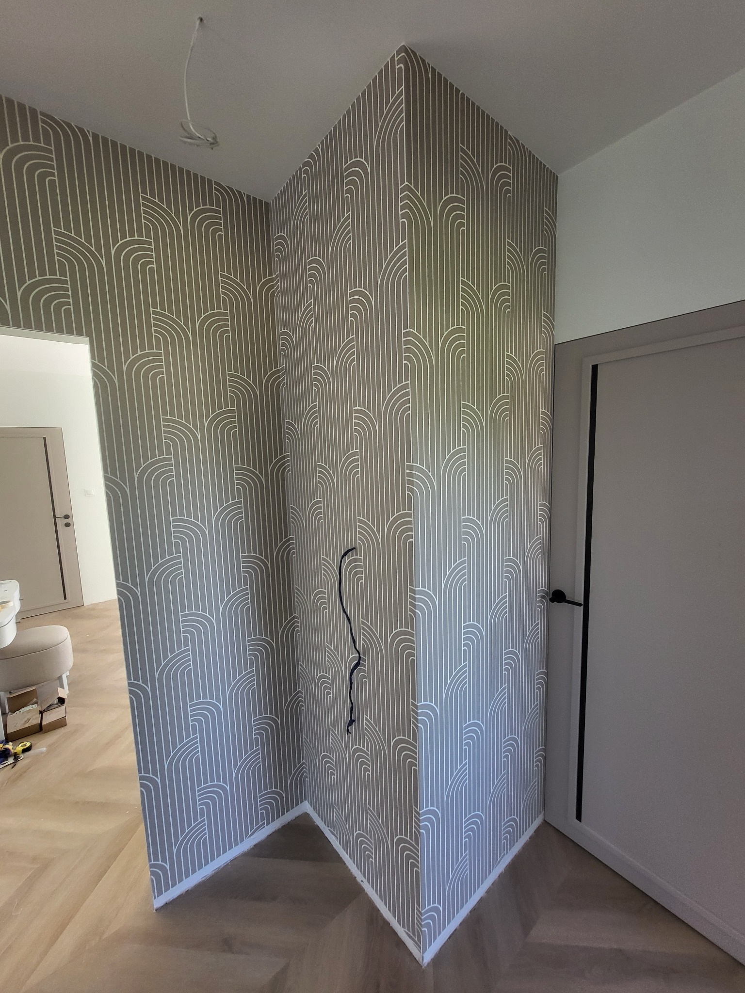 Ściana wykończona tapetą w geometryczny wzór, widoczne narzędzia i fragment podłogi w trakcie prac wykończeniowych. Szare drzwi po prawej stronie.