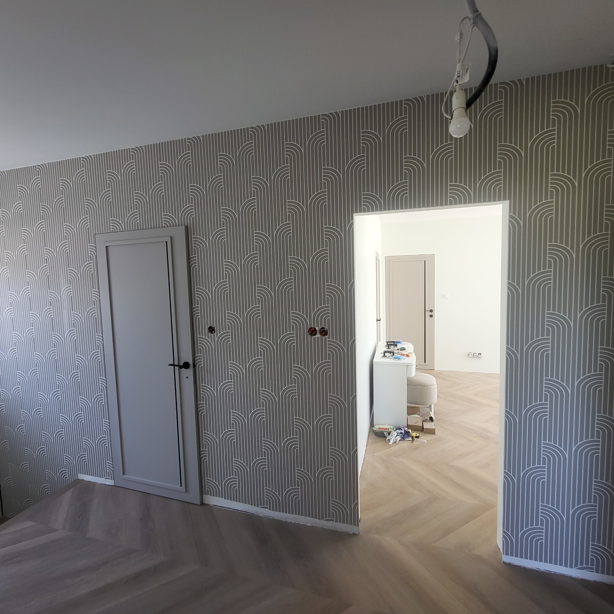 Pokój po remoncie: ściany w szaro-białe wzory, parkiet w jodełkę, drzwi i widok na kolejne pomieszczenie. Widoczne gniazdka elektryczne bez osprzętu.