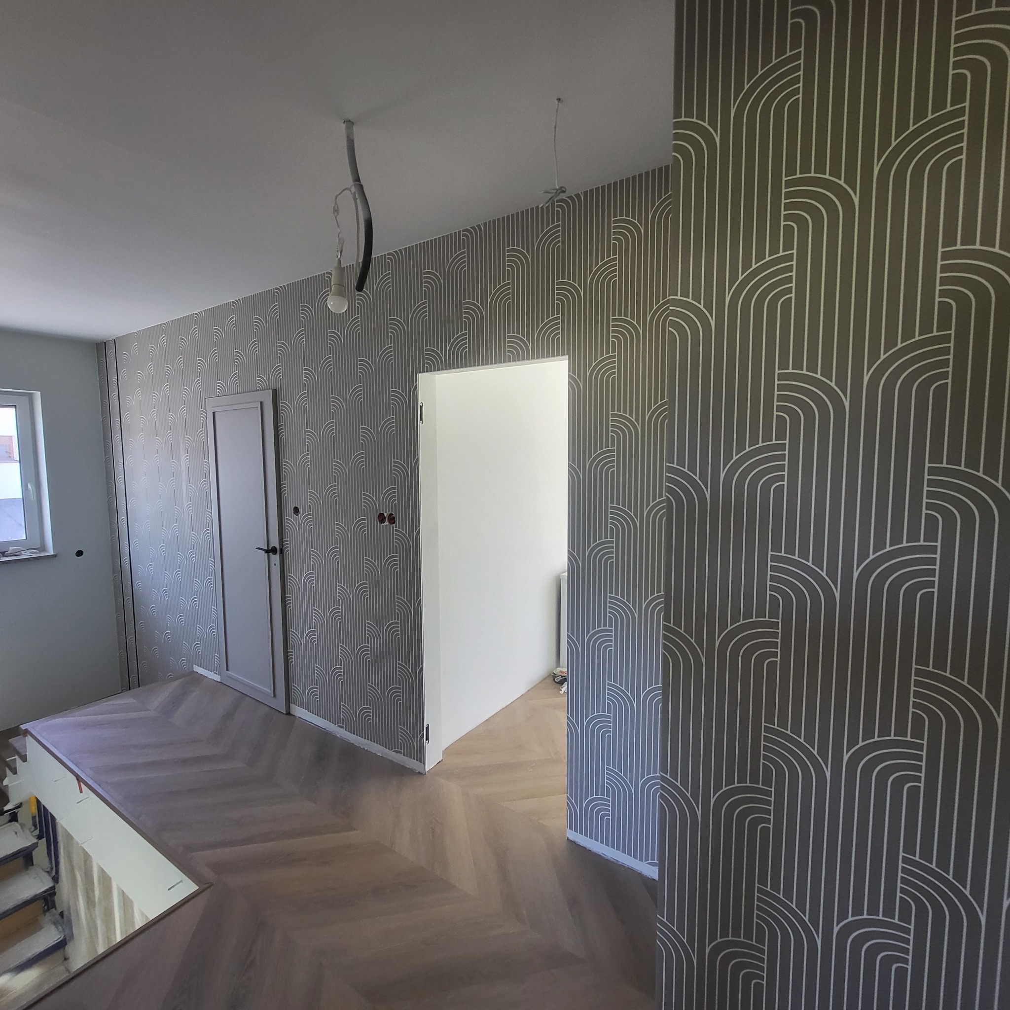 Wykończone wnętrze z wzorzystą tapetą w stylu art déco, parkietem w jodełkę i białymi drzwiami. Widoczne niedokończone instalacje elektryczne.