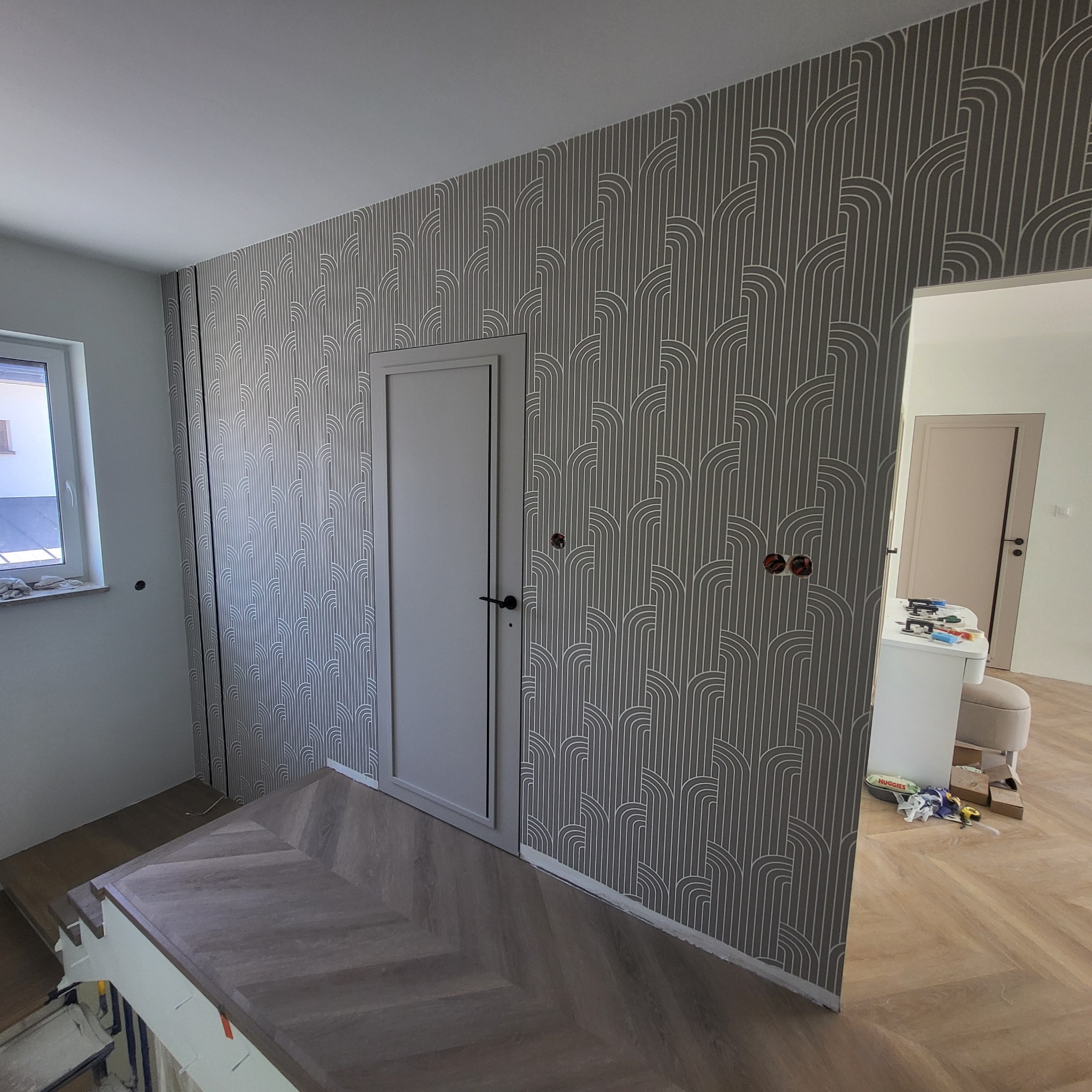 Korytarz po tapetowaniu ścian w szaro-białe wzory art deco. Widoczne drzwi, fragment okna i podłoga ułożona w jodełkę. Świeżo położona tapeta z drobnymi niedoróbkami.