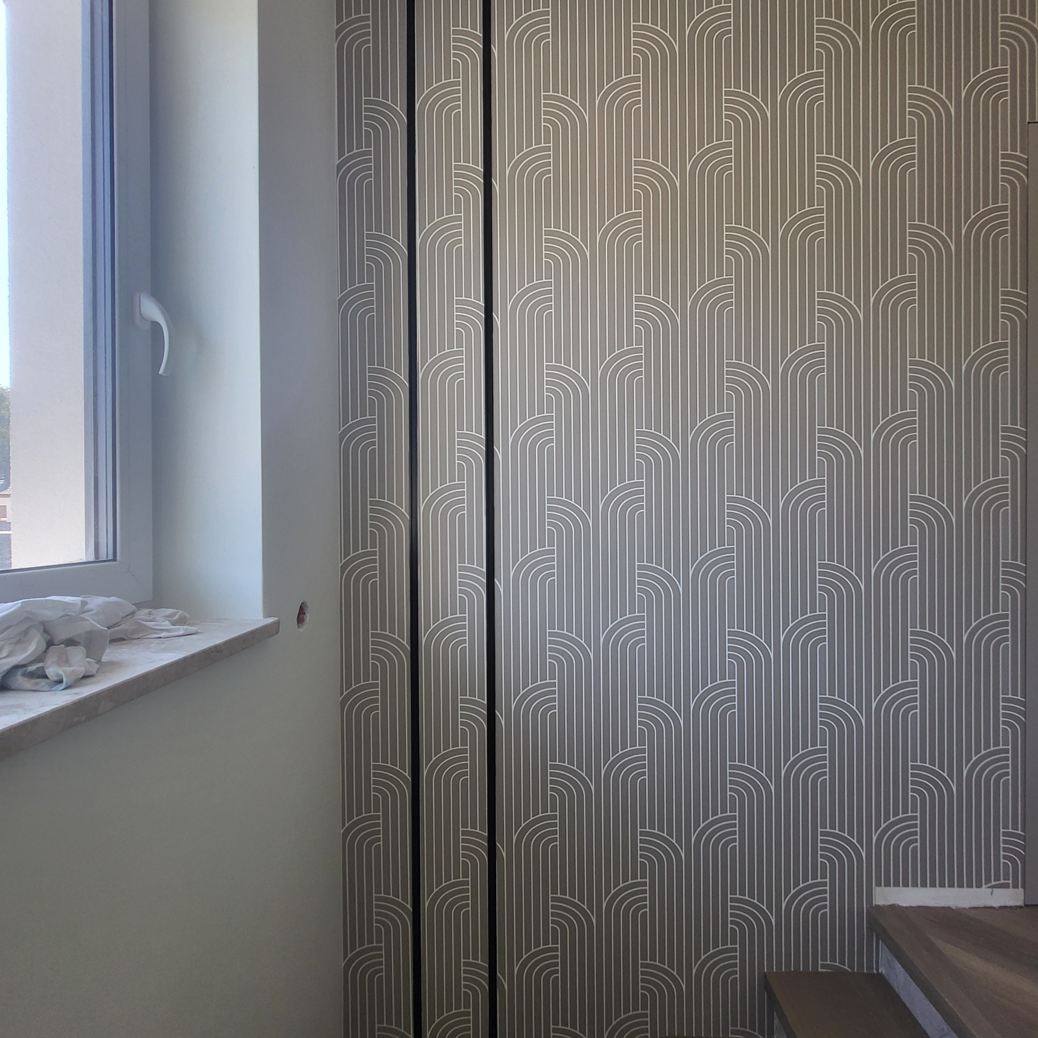 Ściana z szarą tapetą w geometryczne wzory, fragment okna z marmurowym parapetem i schody. Widoczne niedoskonałości przy podłodze.