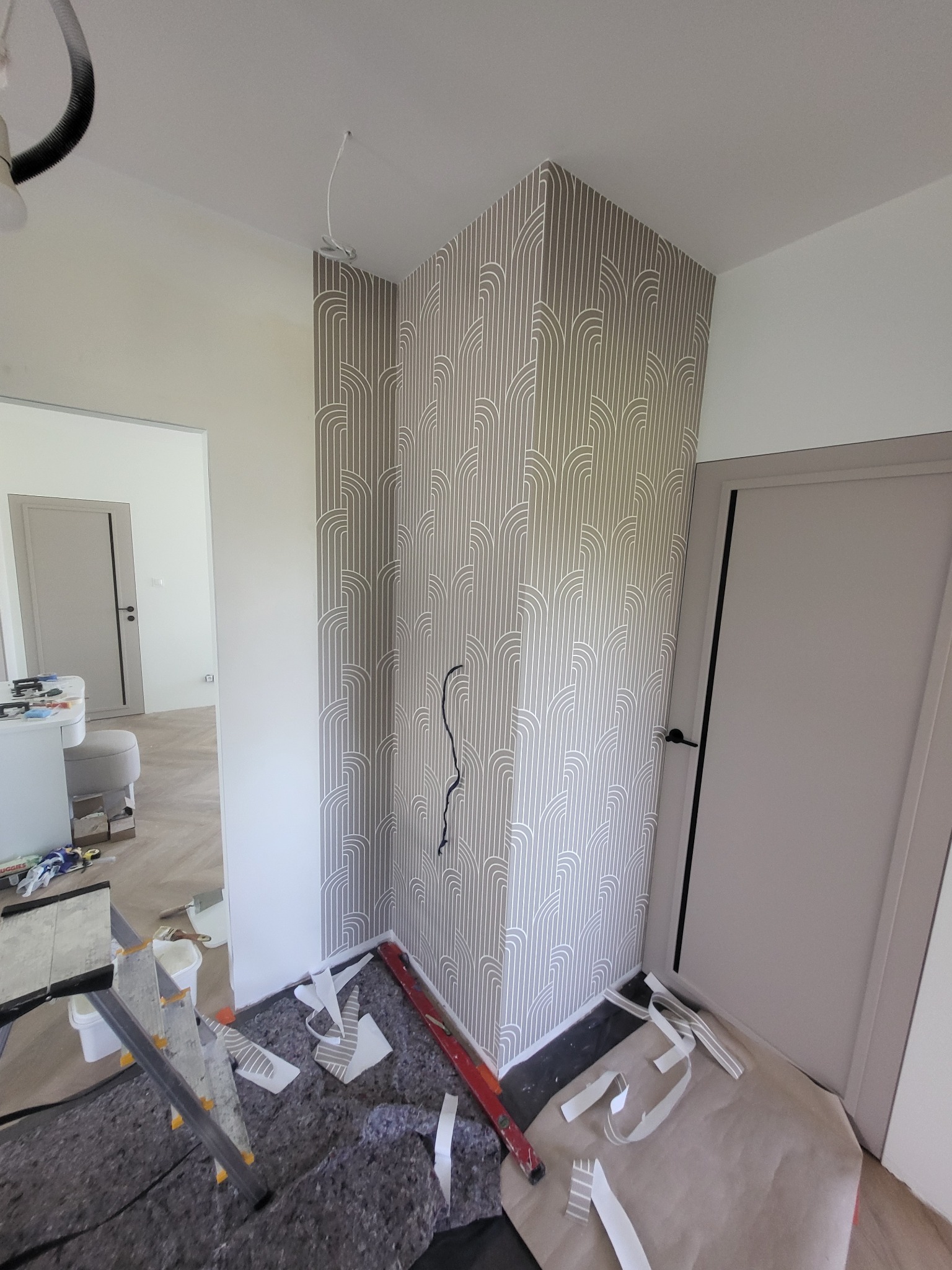 Nakładanie wzorzystej tapety na ścianę w rogu pokoju, widoczne narzędzia i resztki tapety na podłodze, szara kolorystyka, kabel wystający ze ściany.