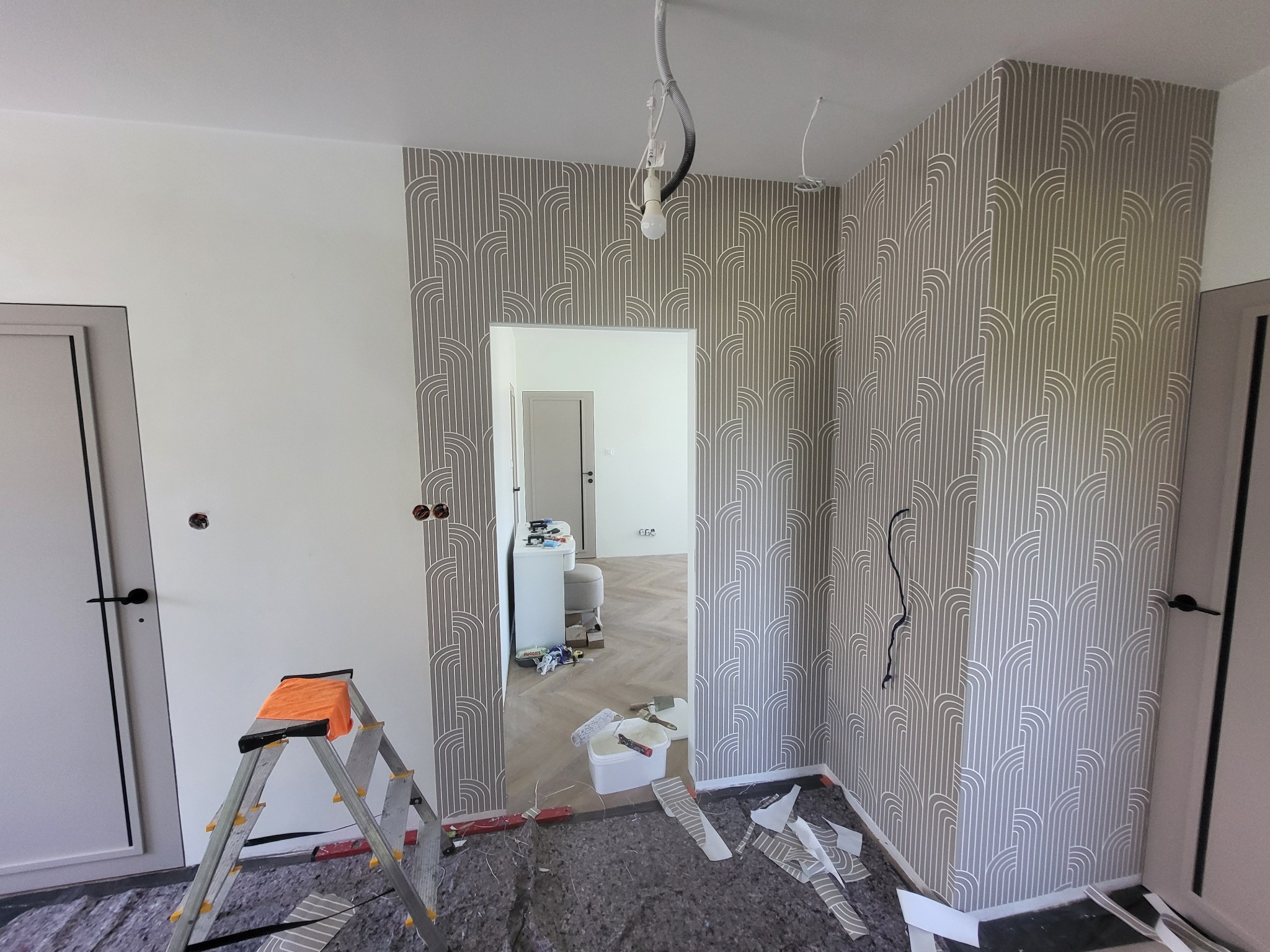 Wnętrze po tapetowaniu ścian w geometryczny wzór. Widoczna drabina, wiadro z klejem i narzędzia malarskie. Kabel zwisający z sufitu.