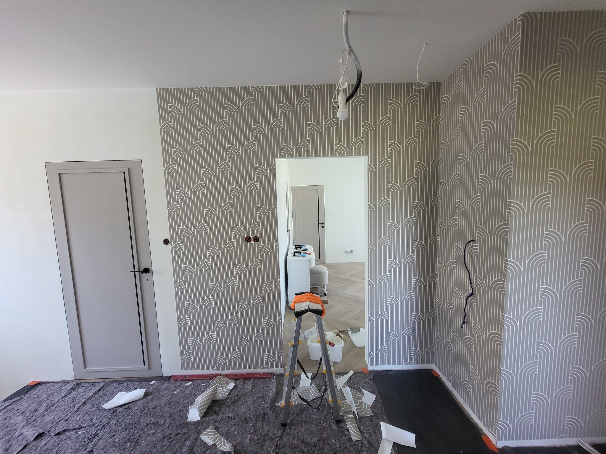 Pokój po tapetowaniu w szaro-białe wzory. Widoczna drabina, fragment podłogi zabezpieczony folią i drzwi w kolorze szarym. Wiszące kable od oświetlenia.