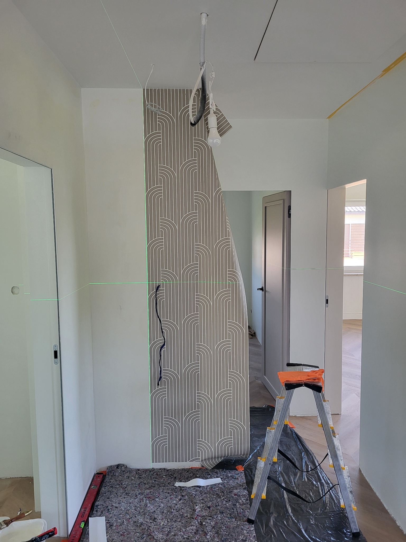 Nakładanie tapety w korytarzu z użyciem poziomicy laserowej. Fragment tapety z geometrycznym wzorem przyklejony do ściany. Widoczna drabina i narzędzia.
