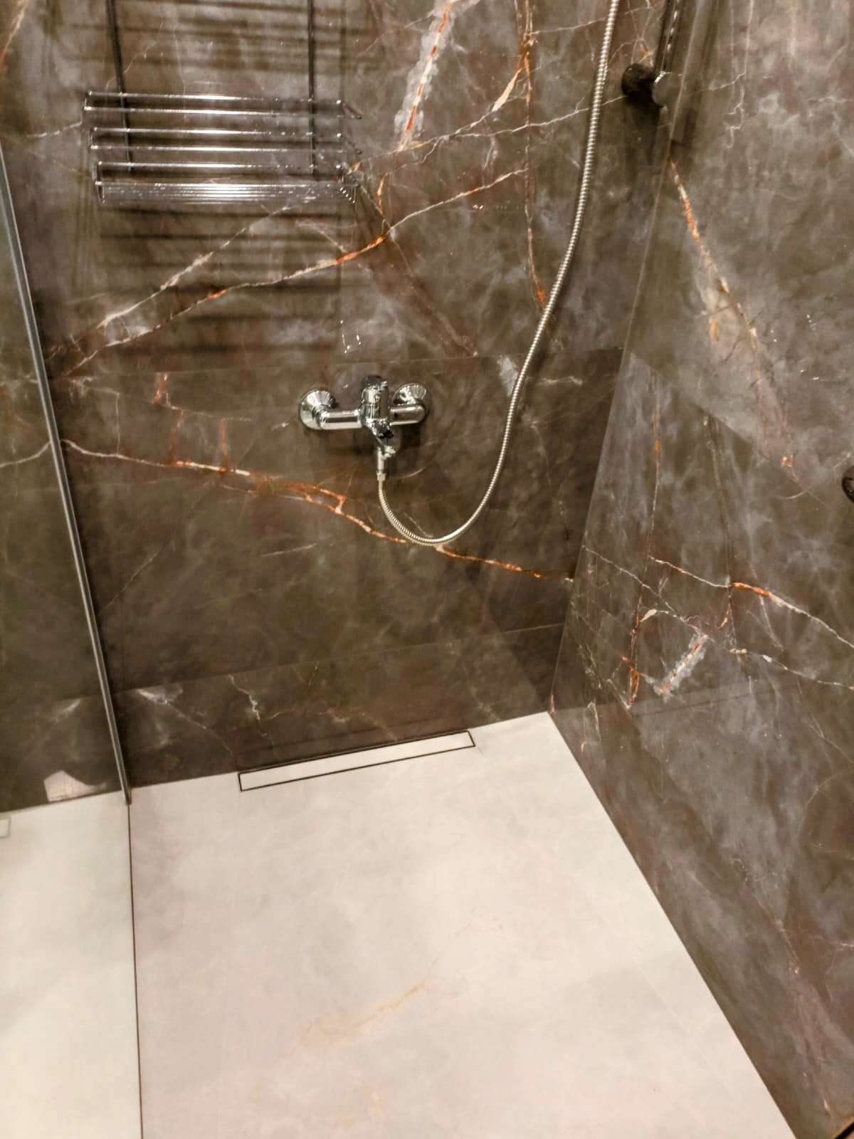 Nowoczesna kabina prysznicowa z marmurowymi płytkami w odcieniach brązu i beżu, chromowana armatura, odpływ liniowy. Eleganckie wykończenie.