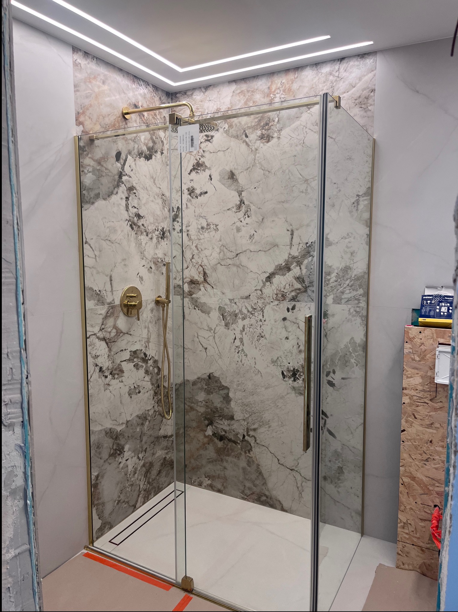 Kabina prysznicowa z marmurowymi płytkami i złotymi akcentami, oświetlenie LED w suficie. Widoczne detale wykończenia i elementy instalacji.