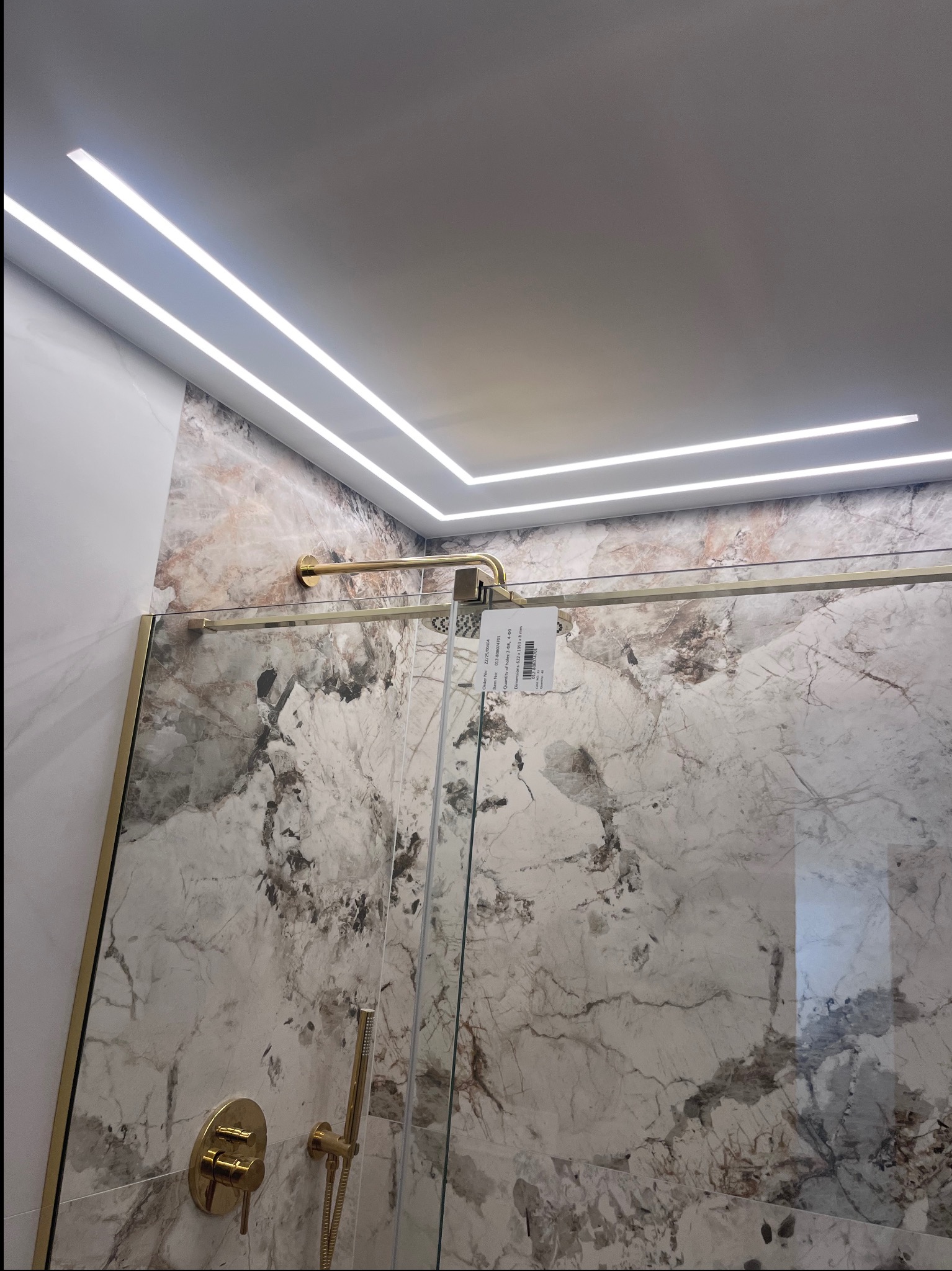 Nowoczesna łazienka z marmurowymi płytkami, złotą armaturą i liniowym oświetleniem LED w suficie. Kabina prysznicowa ze złotymi profilami.