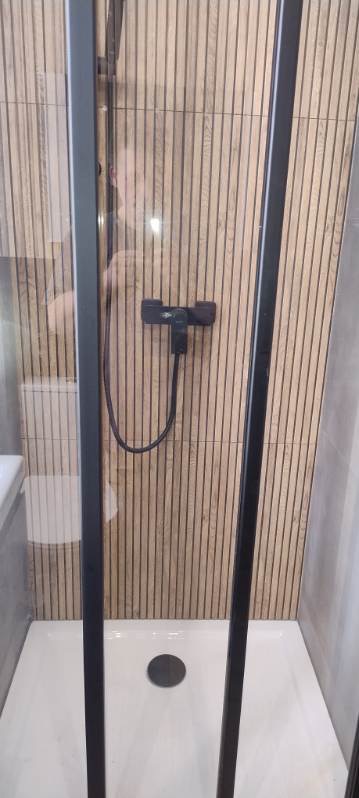 Kabina prysznicowa z czarnymi profilami i drewnopodobnymi płytkami na ścianie. Widoczny biały brodzik i czarna bateria. Minimalistyczny design.