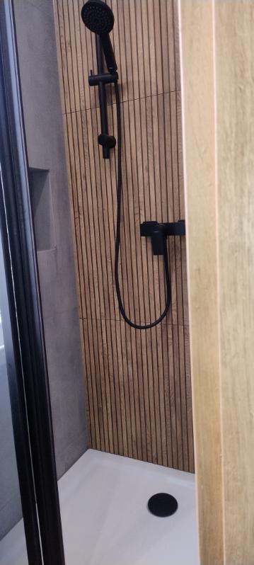 Nowoczesna kabina prysznicowa z czarną armaturą i drewnopodobnymi płytkami ściennymi. Biały brodzik z odpływem punktowym. Minimalistyczny design.