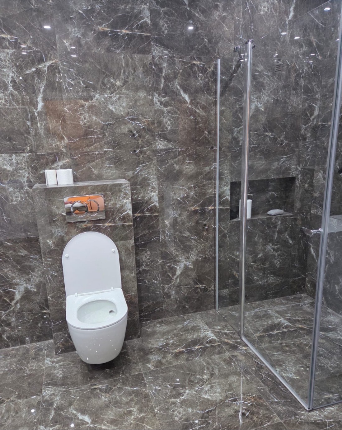 Nowoczesna łazienka z kabiną prysznicową i toaletą, wykończona płytkami imitującymi marmur w odcieniach szarości. Minimalistyczny design, oświetlenie punktowe.