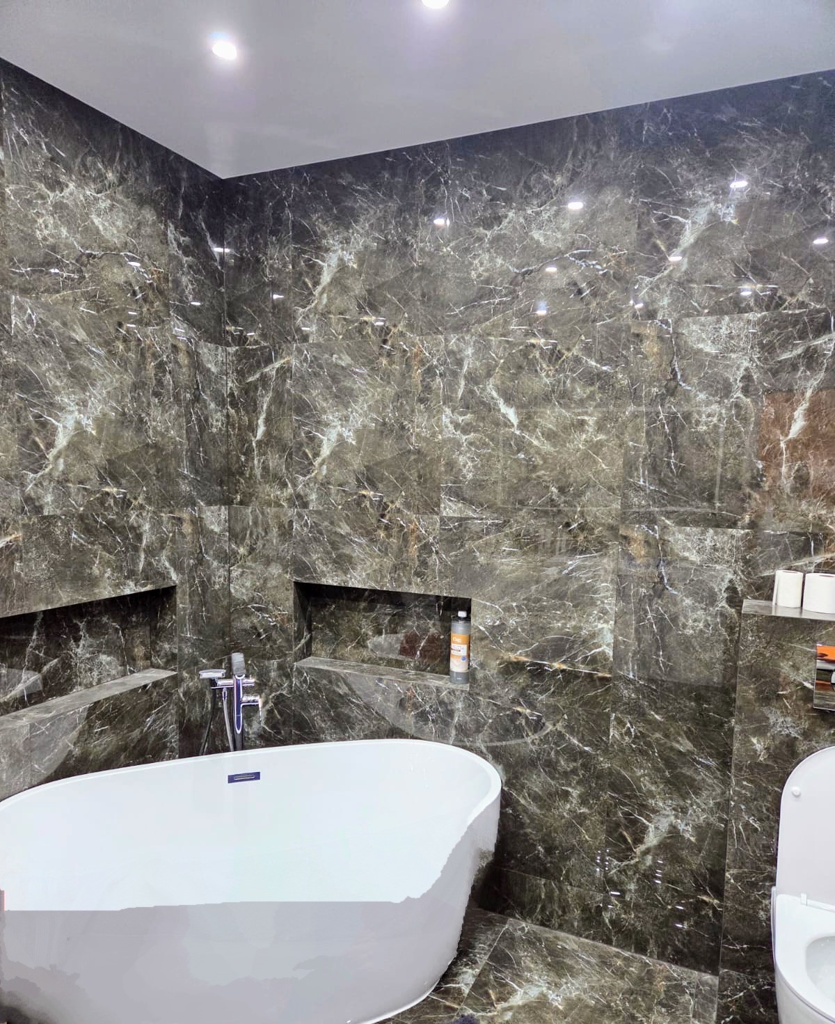 Elegancka łazienka z wanną wolnostojącą i ciemnymi płytkami imitującymi marmur, z wnękami na kosmetyki i akcesoria, oświetlona punktowo.