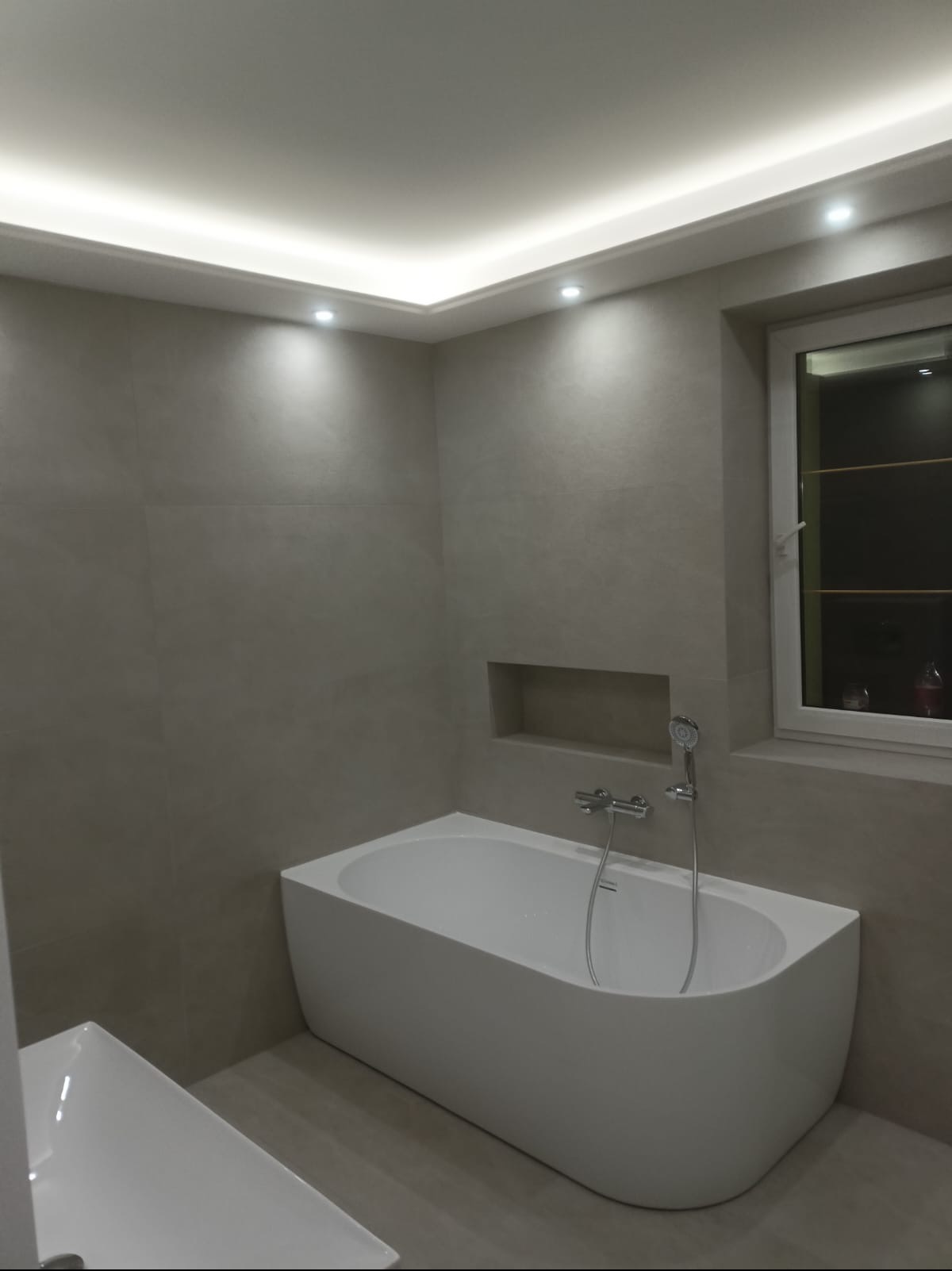 Nowoczesna łazienka w szarościach z białą wanną, oświetleniem LED i wnęką w ścianie. Minimalistyczny design, widok na okno z półkami. Wykończenie z dbałością o detale.