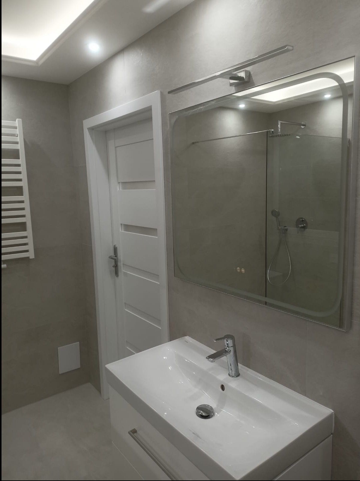 Nowoczesna łazienka w odcieniach szarości z białą umywalką, lustrem z oświetleniem i grzejnikiem drabinkowym. Widoczna kabina prysznicowa w odbiciu lustra.