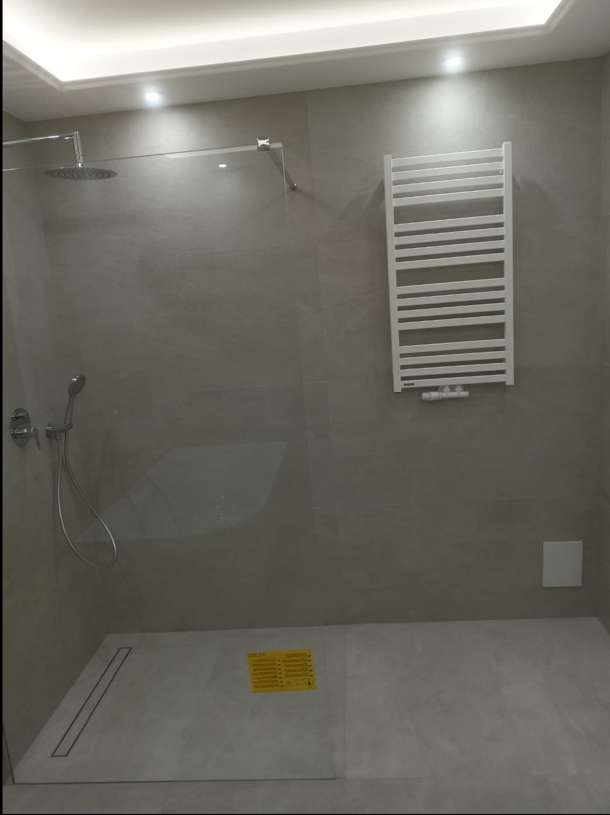 Nowoczesna łazienka z kabiną prysznicową typu walk-in, grzejnikiem drabinkowym i szarymi płytkami. Minimalistyczny design, oświetlenie punktowe.