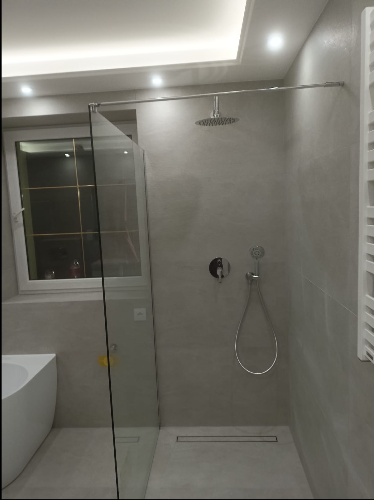 Nowoczesna kabina prysznicowa typu walk-in z deszczownicą i odpływem liniowym, wykończona szarą płytką. Widoczne okno z szybą i fragment wanny.