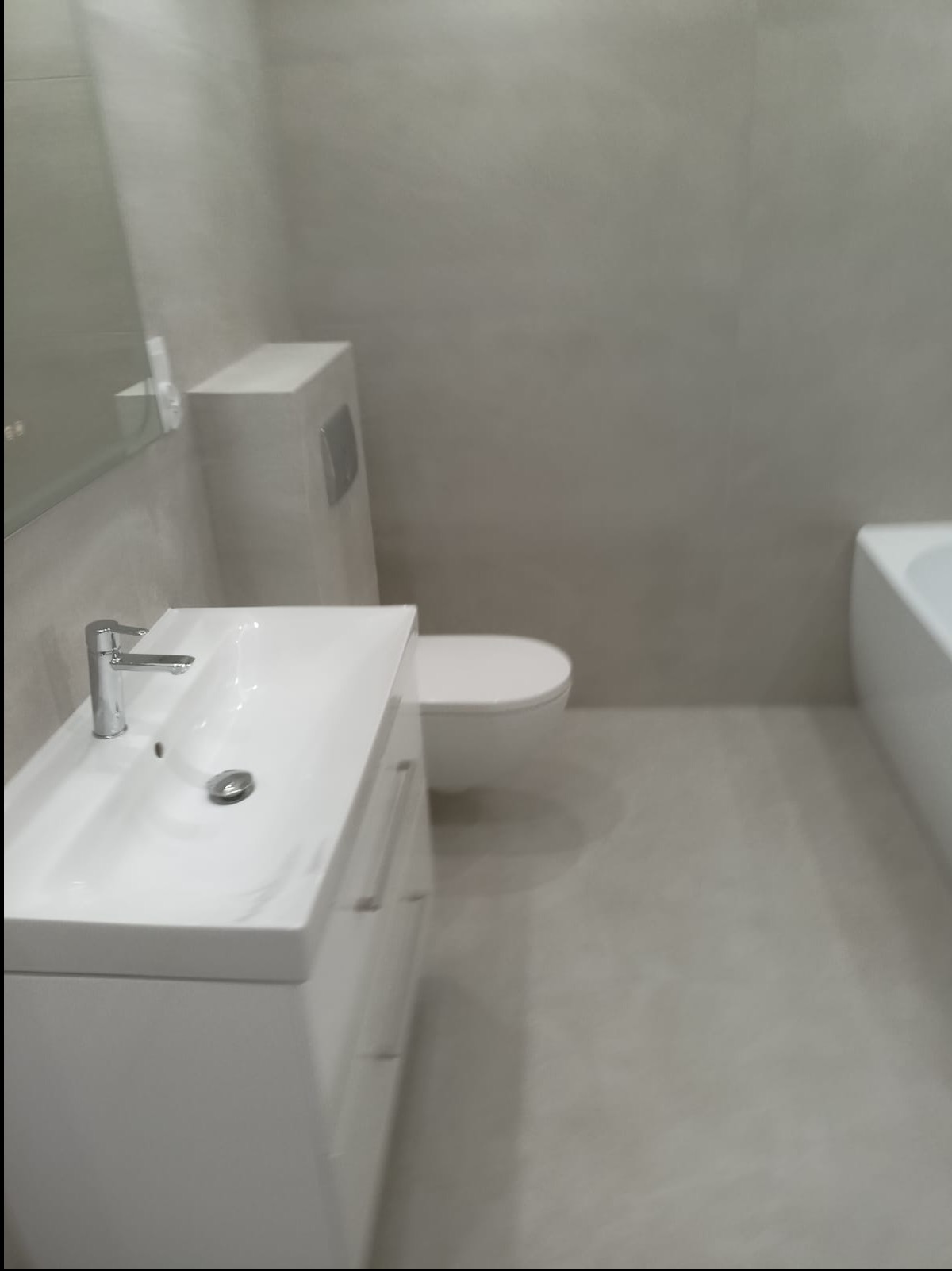 Jasna łazienka po remoncie: biała umywalka z szafką, toaleta i fragment wanny na tle szarych płytek. Minimalistyczny design, nowoczesne wykończenie.