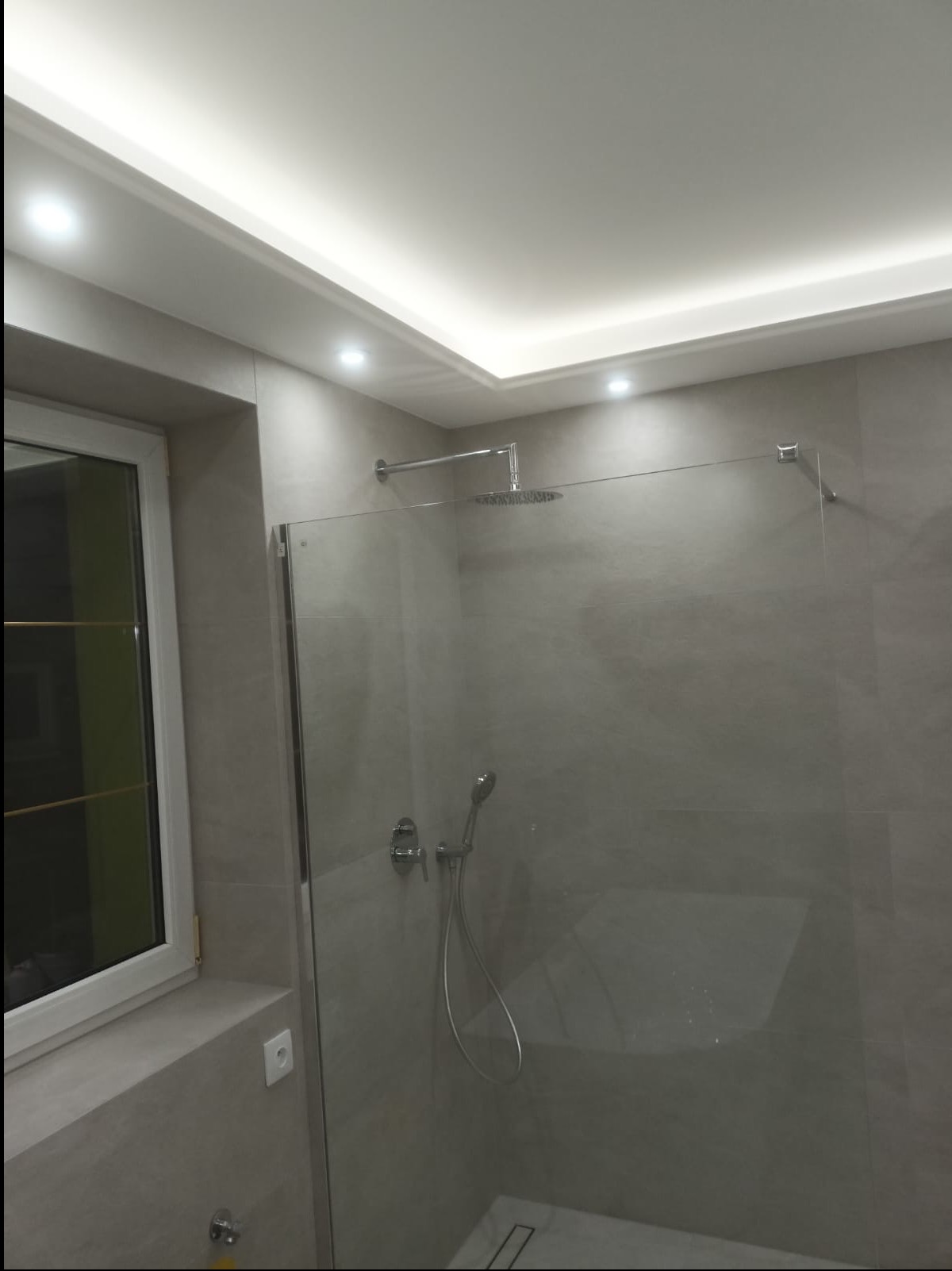 Nowoczesna kabina prysznicowa z deszczownicą i szklaną ścianką, wykończona szarą płytką. Oświetlenie sufitowe LED. Minimalistyczny design.