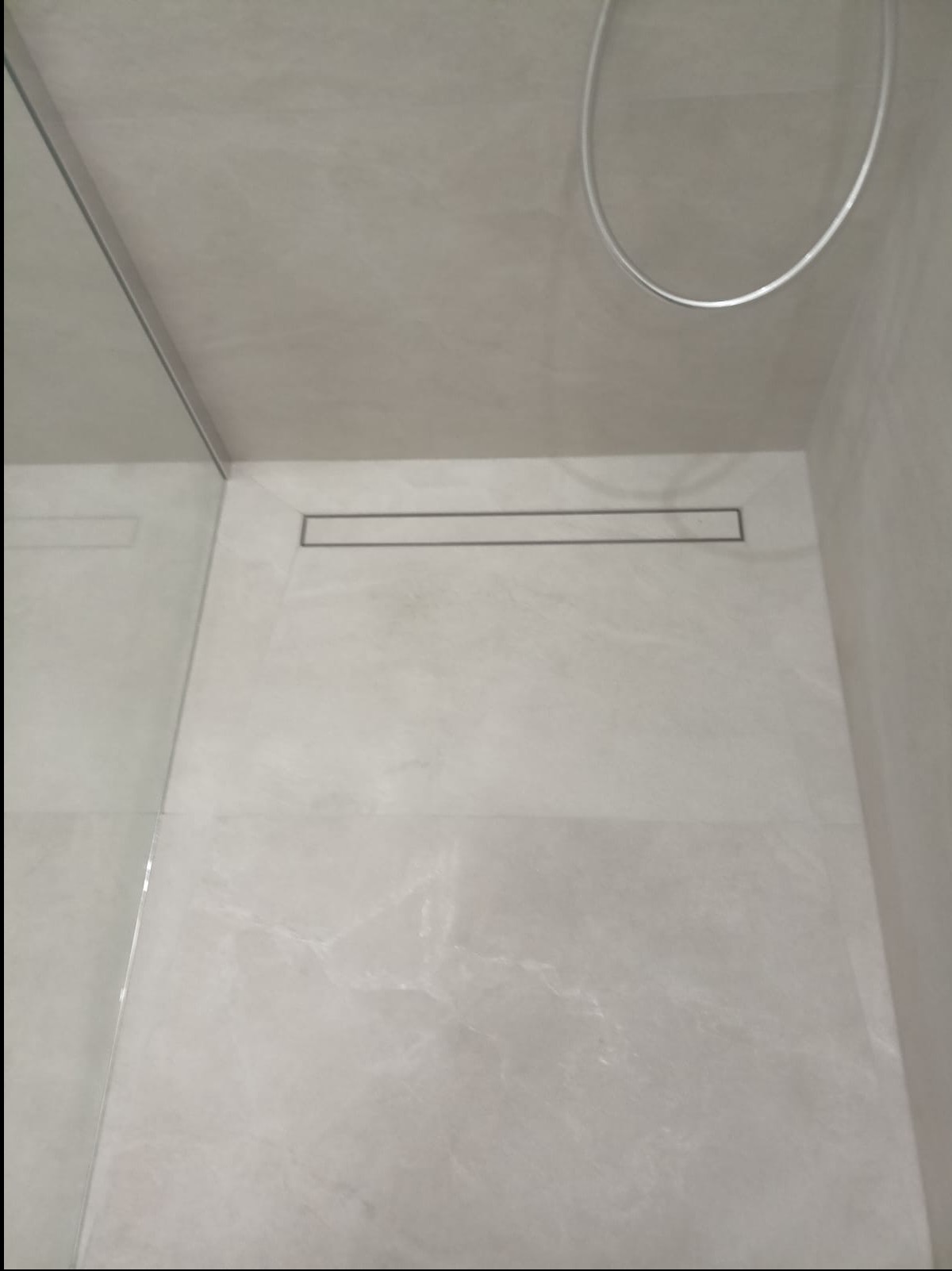 Nowoczesna łazienka z odpływem liniowym w podłodze, wykończona jasnymi płytkami imitującymi marmur. Widoczna szklana ścianka kabiny prysznicowej i chromowana deszczownica.