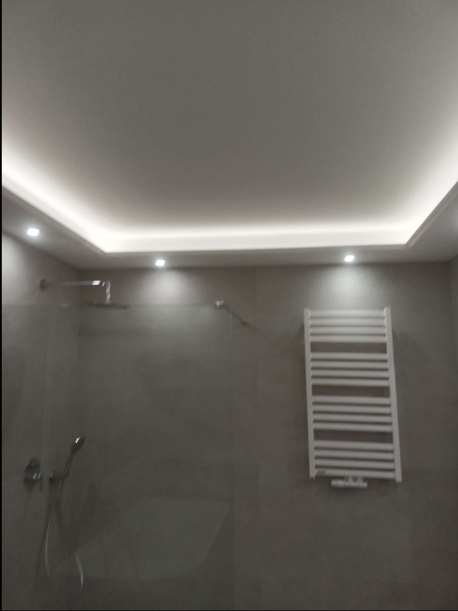 Nowoczesna łazienka z szarymi płytkami, oświetleniem LED na suficie i białym grzejnikiem drabinkowym. Minimalistyczny design, kabina prysznicowa ze szklaną ścianką.