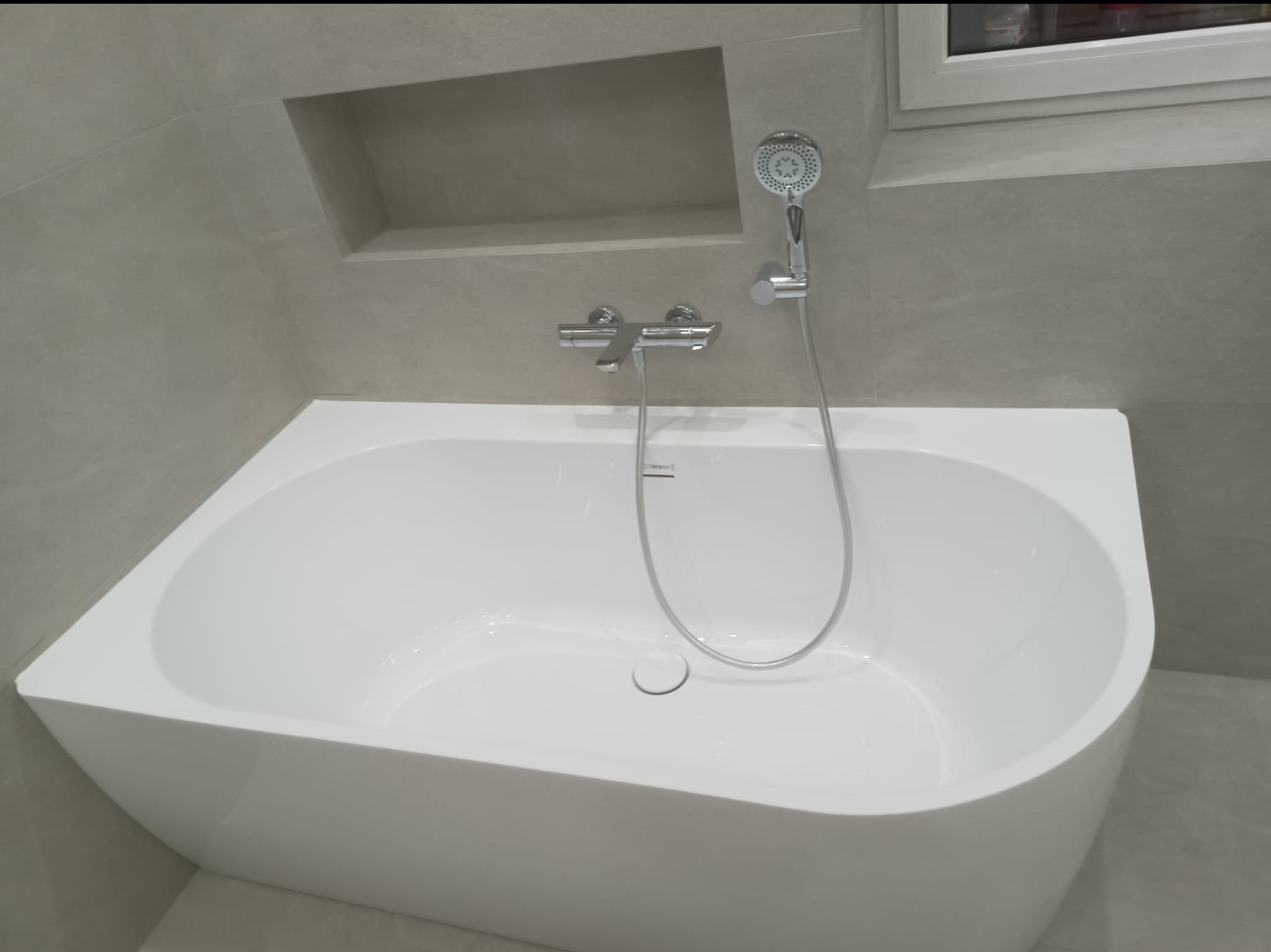 Biała wanna z baterią i słuchawką prysznicową na tle szarych płytek. Nad wanną wnęka w ścianie. Minimalistyczny design łazienki.