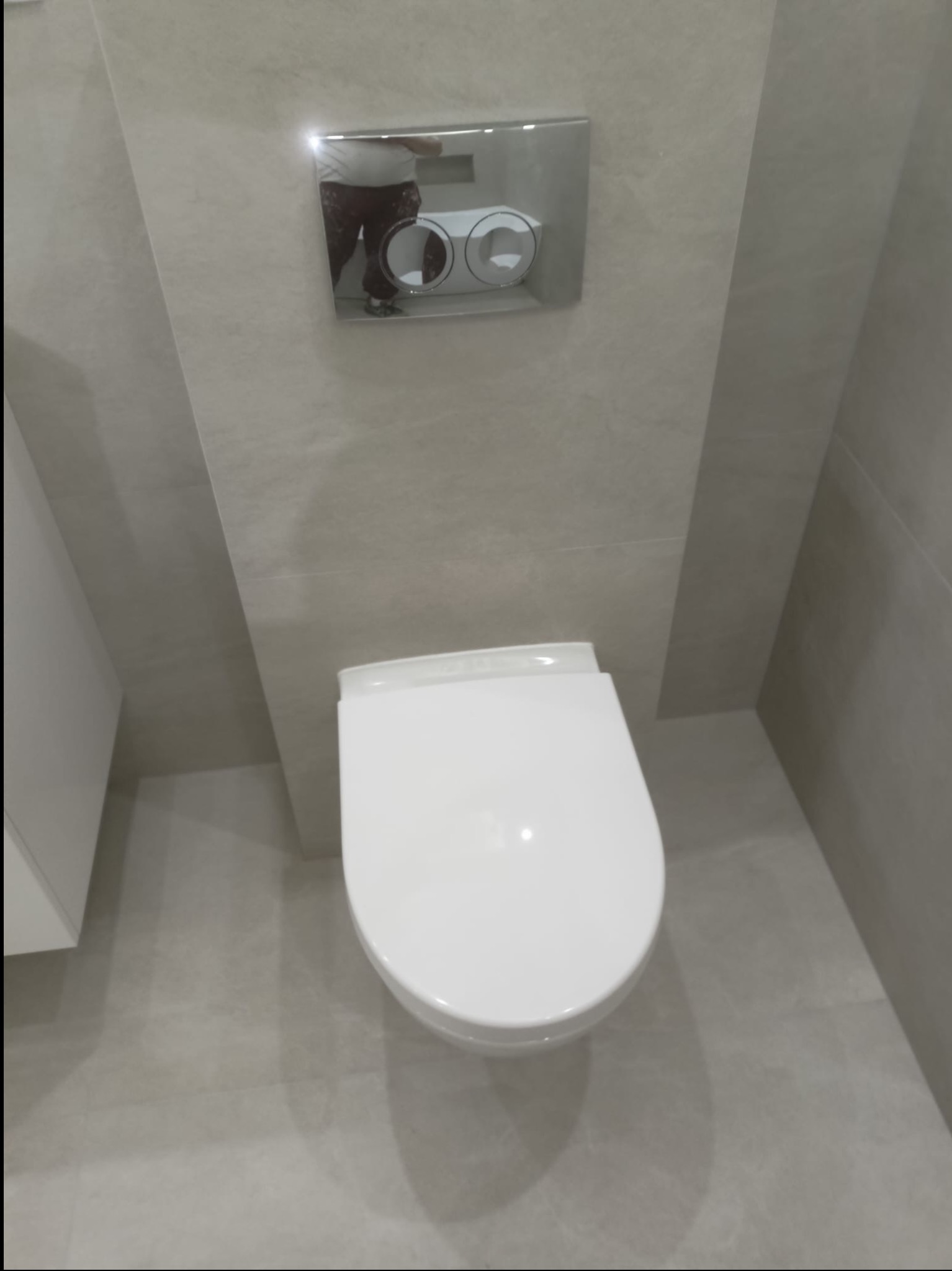 Nowoczesna toaleta podwieszana z chromowanym przyciskiem spłukującym, zamontowana na ścianie wyłożonej jasnymi, matowymi płytkami w minimalistycznej łazience.