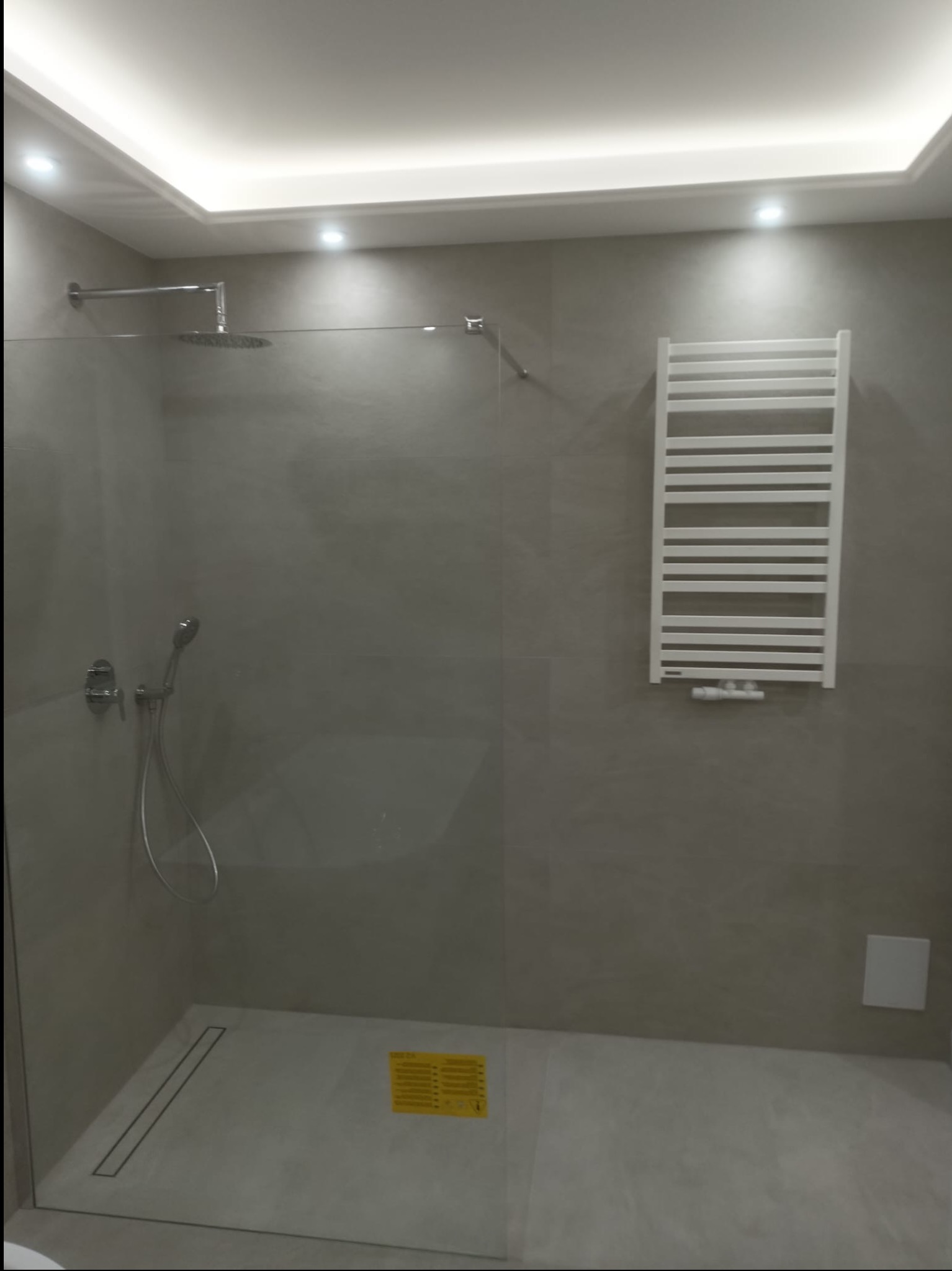 Nowoczesna kabina prysznicowa walk-in z odpływem liniowym i grzejnikiem drabinkowym na szarej ścianie. Oświetlenie sufitowe LED.