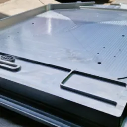 Precyzyjnie obrobiony, aluminiowy element z frezowanymi wzorami i wgłębieniami. Widoczne ślady obróbki skrawaniem i logo producenta. Detal gotowy do użycia.