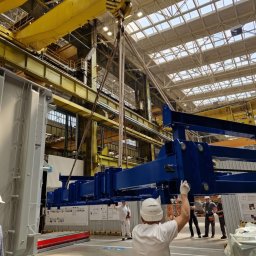 FERROTECH SPÓŁKA CYWILNA - Fabryka: suwnica podnosi niebieską konstrukcję stalową. Pracownicy w kaskach nadzorują proces. Żółte elementy konstrukcji hali w tle.