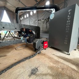 FERROTECH SPÓŁKA CYWILNA - Spawanie konstrukcji stalowej w hali namiotowej. Widoczny spawacz w masce, urządzenie spawalnicze Lincoln i filtr powietrza Filtrobox 1500.