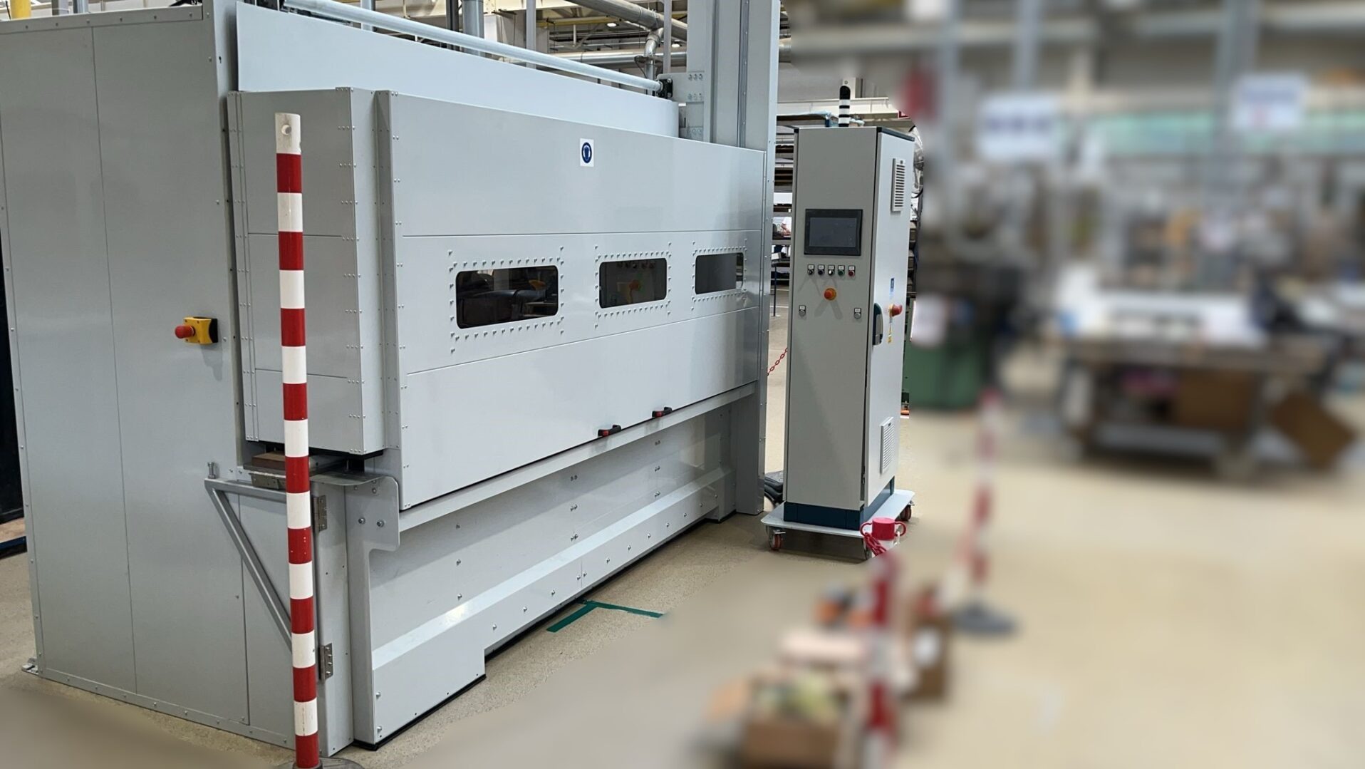 Duża, szara maszyna przemysłowa z panelem sterowania na kółkach, widoczna w hali produkcyjnej z rozmytym tłem.