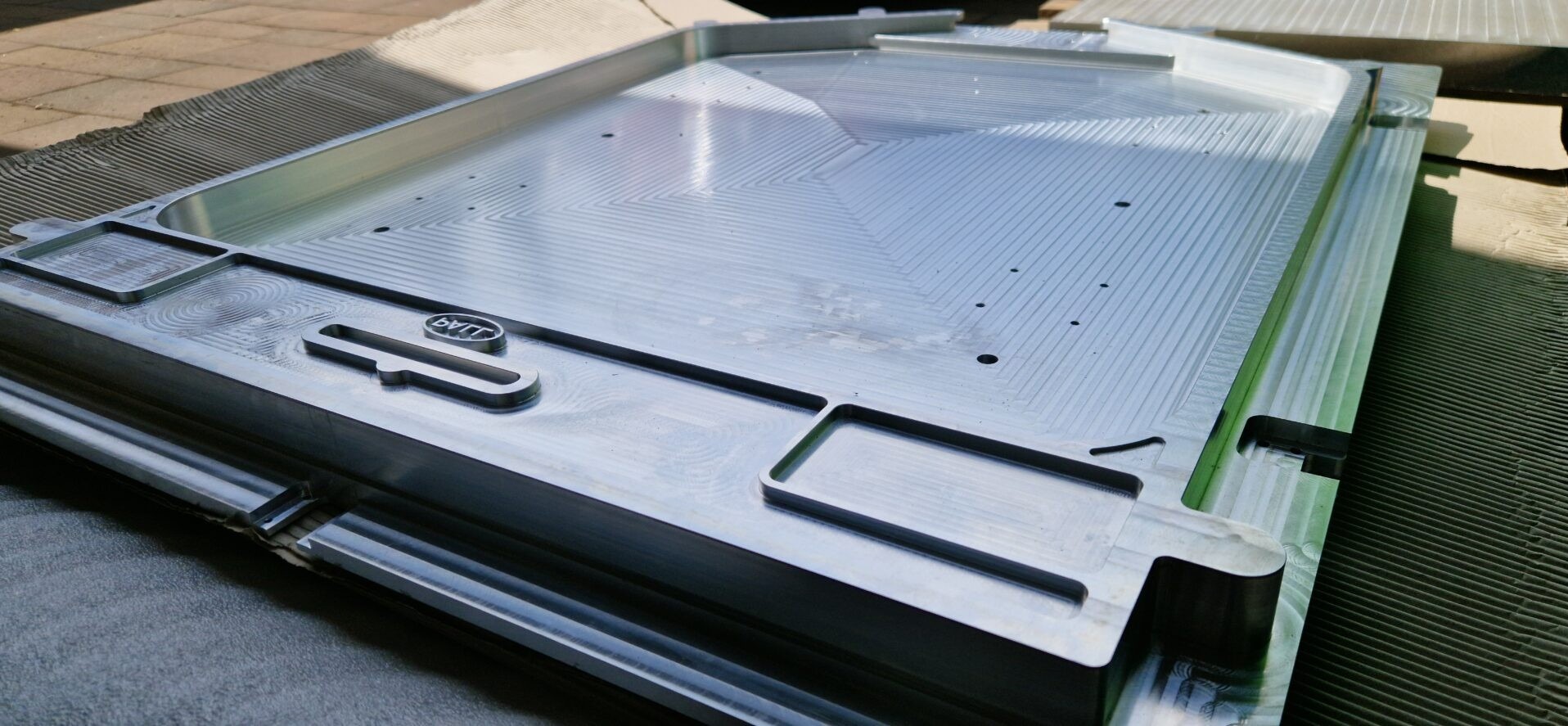 Precyzyjnie obrobiony, aluminiowy element z frezowanymi wzorami i wgłębieniami. Widoczne ślady obróbki skrawaniem i logo producenta. Detal gotowy do użycia.