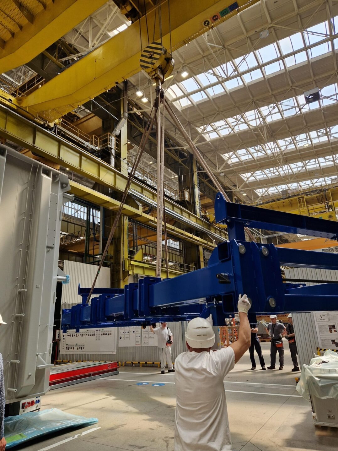 Fabryka: suwnica podnosi niebieską konstrukcję stalową. Pracownicy w kaskach nadzorują proces. Żółte elementy konstrukcji hali w tle.