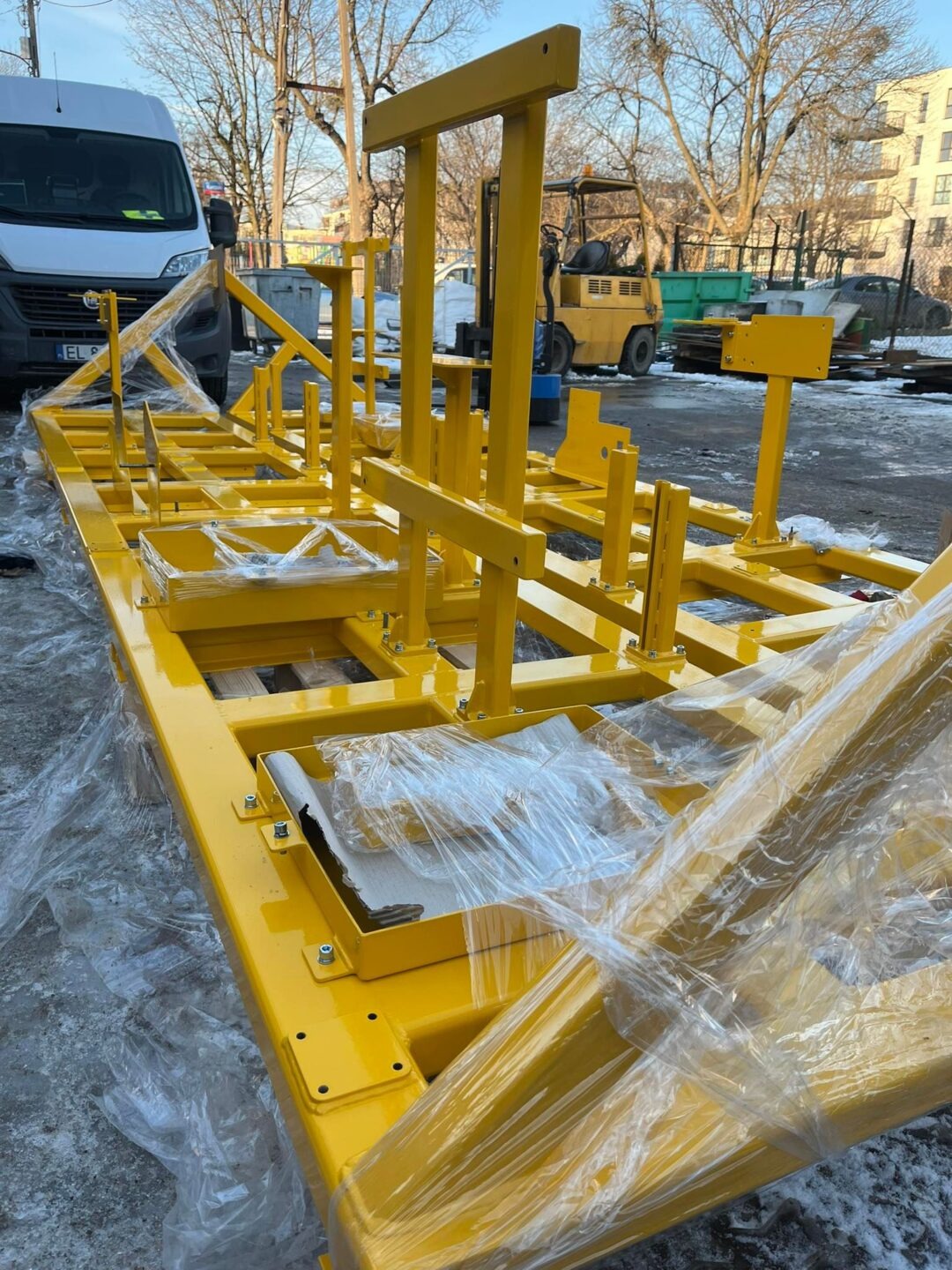 Żółta, metalowa konstrukcja nośna, owinięta folią ochronną, na zewnątrz. W tle widoczny wózek widłowy i samochód dostawczy. Konstrukcja spawana i pomalowana.