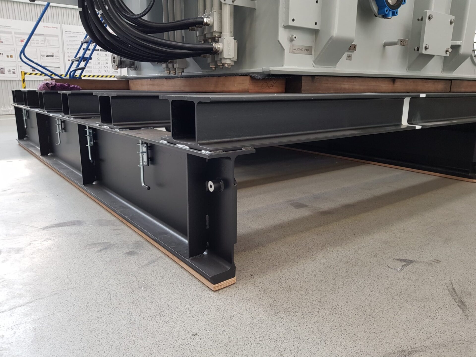 Czarna, spawana konstrukcja stalowa z belek H, podtrzymująca duży, szary transformator. Widoczne kable i tabliczka znamionowa 'JACKING PAD'.