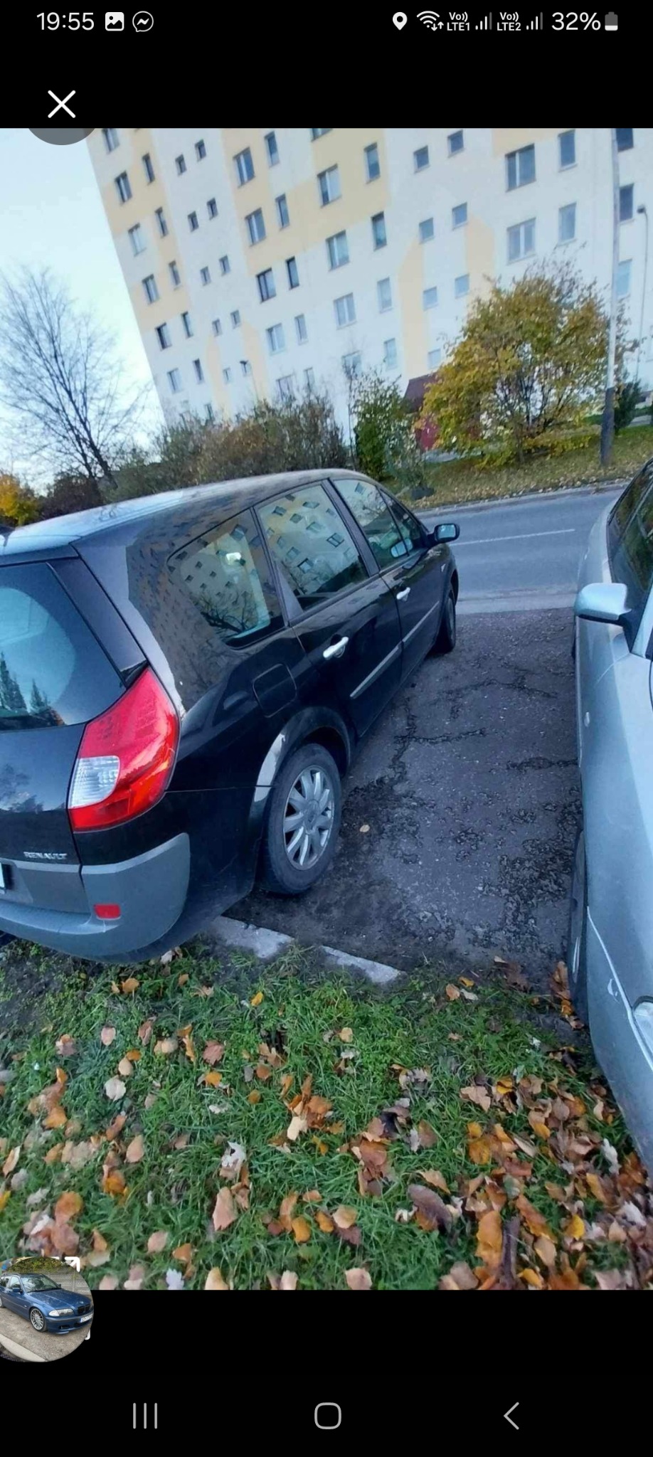 Czarny samochód Renault Scenic zaparkowany na nierównej nawierzchni, widok z tyłu, w tle budynek mieszkalny i drzewa. Jesienna sceneria.