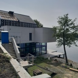 JANUSZ OMYŁA FIRMA USŁUGOWO - HANDLOWA - Nowoczesny dom murowany z płaskim dachem w trakcie budowy, widok z zewnątrz na częściowo wykończoną elewację, schody i pomost nad wodą. Na dachu widoczni robotnicy.