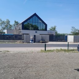 JANUSZ OMYŁA FIRMA USŁUGOWO - HANDLOWA - Nowoczesny dom murowany z dużym przeszkleniem szczytowym i czarną stolarką, widoczny od frontu, z osobą przechodzącą obok ogrodzenia. Częściowo widoczne rusztowania.