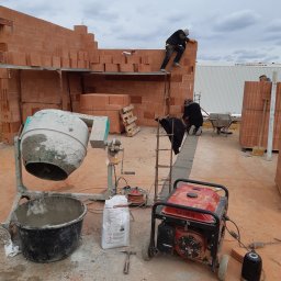 JANUSZ OMYŁA FIRMA USŁUGOWO - HANDLOWA - Budowa domu z czerwonej cegły: murarze pracują na rusztowaniu, widoczna betoniarka, generator prądu i materiały budowlane. Etap wznoszenia ścian.