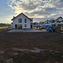 JANUSZ OMYŁA FIRMA USŁUGOWO - HANDLOWA - Nowoczesny dom murowany z płaskim dachem w trakcie budowy, widoczne przygotowane podłoże pod taras i pracownicy. W tle błękitne niebo z chmurami.